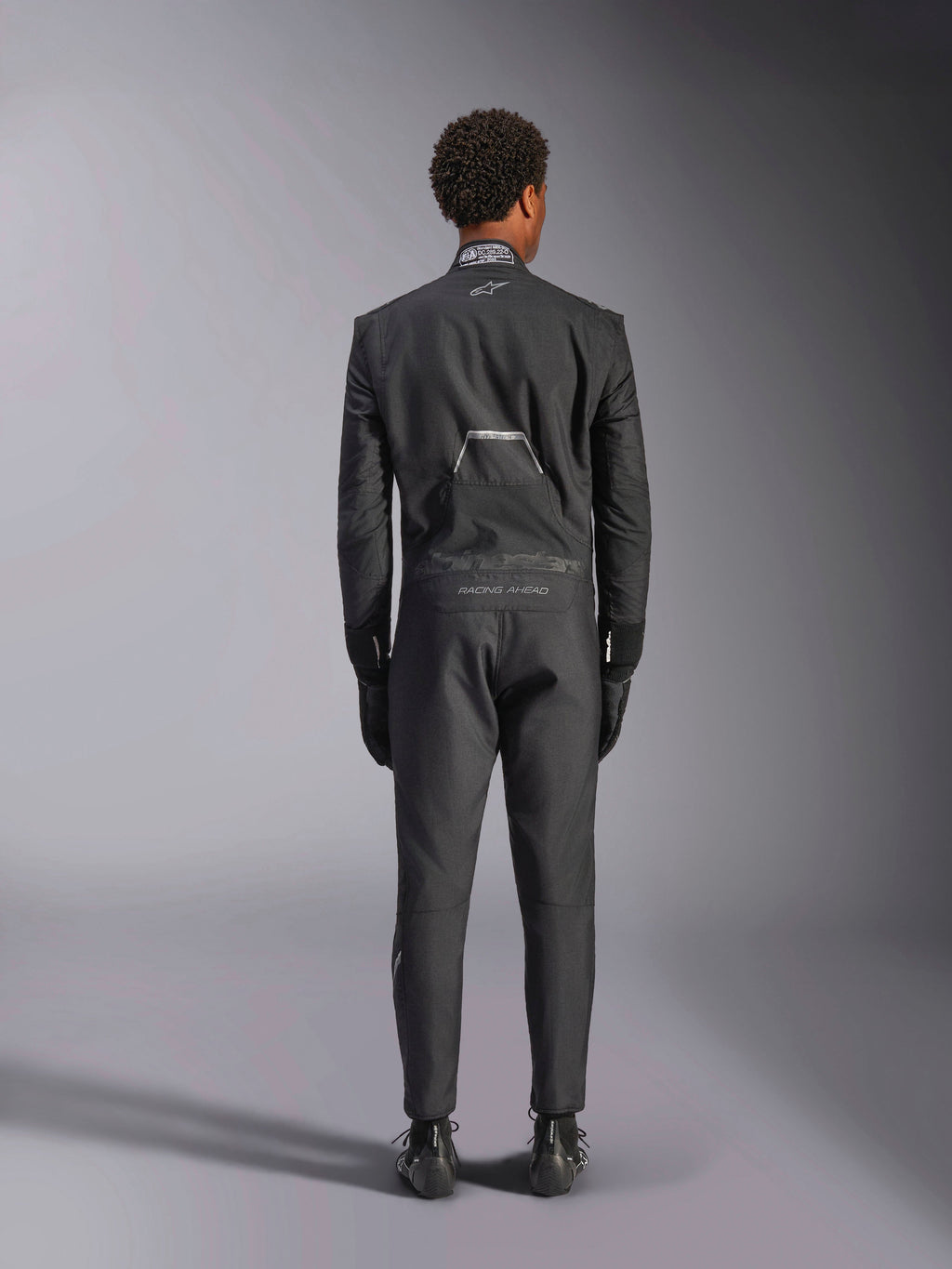 Kombinezon Alpinestars Hypertech V3, Black Dark Shadow, widok z tyłu profesjonalnego jednoczęściowego samochodowego kombinezonu wyścigowego, z etykietą certyfikacji FIA na kołnierzu oraz brandingiem „Alpinestars” i „RACING AHEAD” na dolnej części pleców, noszony z dopasowanymi czarnymi rękawicami i butami wyścigowymi.