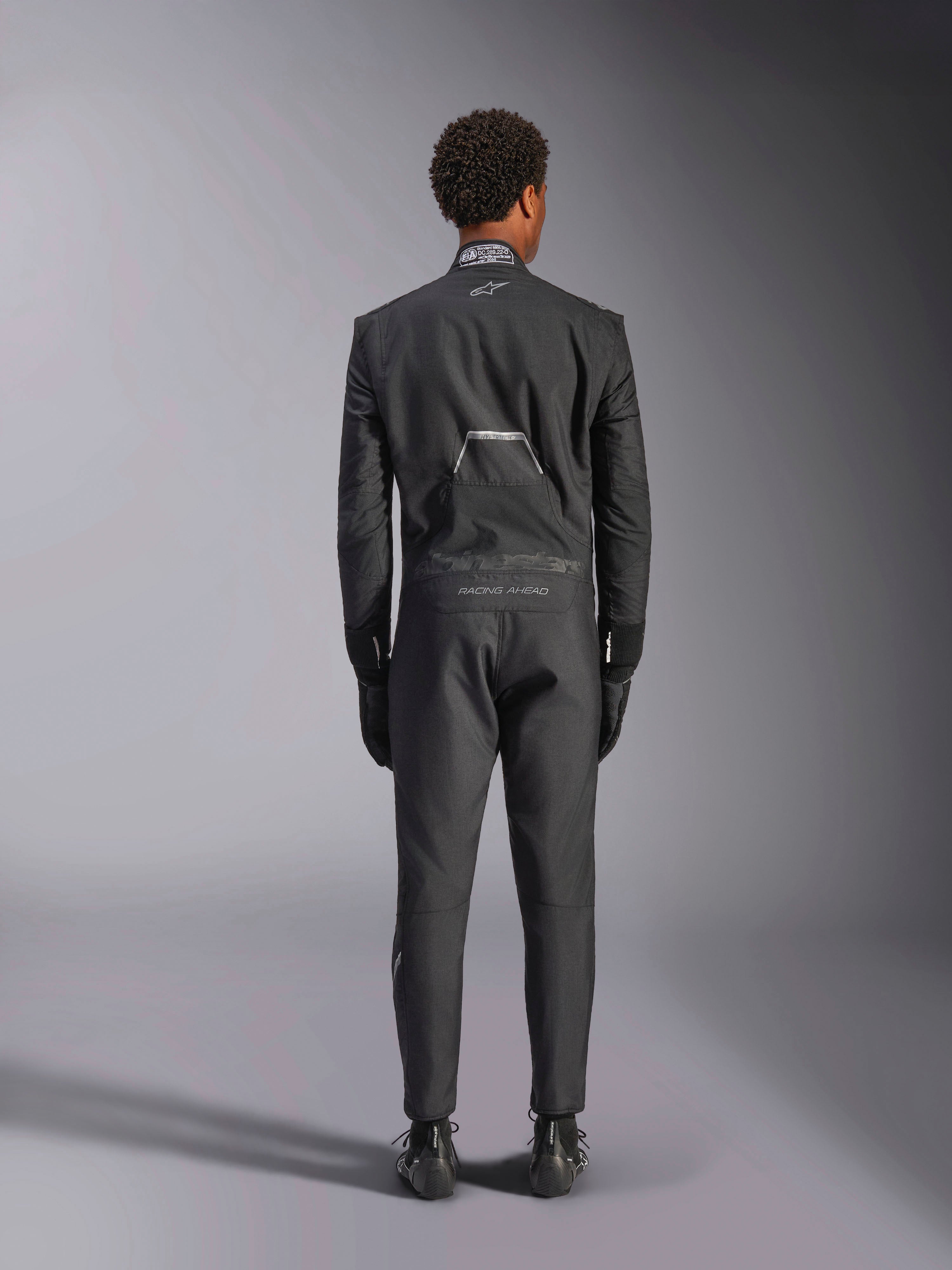 Kombinezon Alpinestars Hypertech V3, Black Dark Shadow, widok z tyłu profesjonalnego jednoczęściowego samochodowego kombinezonu wyścigowego, z etykietą certyfikacji FIA na kołnierzu oraz brandingiem „Alpinestars” i „RACING AHEAD” na dolnej części pleców, noszony z dopasowanymi czarnymi rękawicami i butami wyścigowymi.
