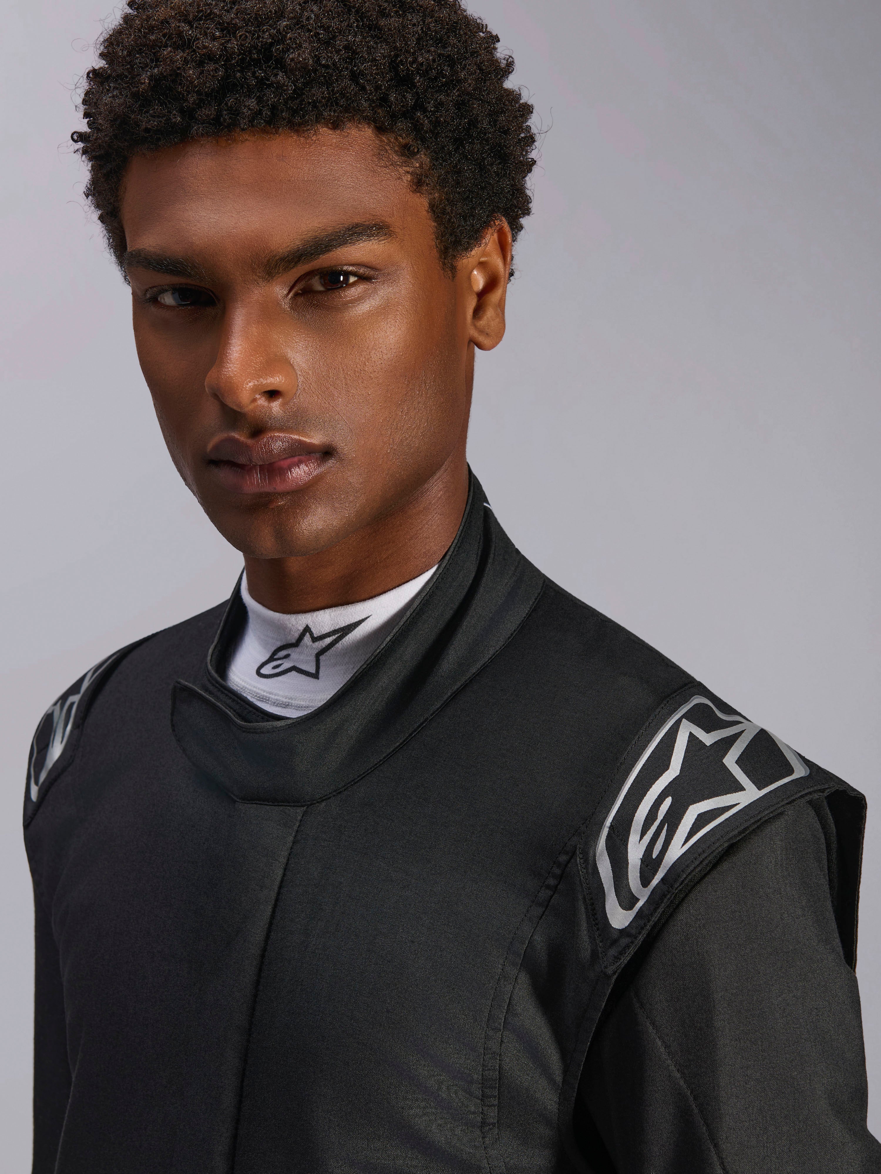 Kombinezon Alpinestars Hypertech V3, samochodowy kombinezon wyścigowy Black Dark Shadow, zbliżenie na modela, srebrne logo na ramionach, wysoki kołnierz z białym półgolfem Alpinestars, profesjonalna odzież ochronna do sportów motorowych