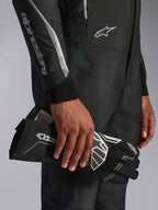 Alpinestars Hypertech V3 Suit, kombinezon w kolorze Black Dark Shadow, zbliżenie na detale rękawa i torsu ze srebrnym logo Alpinestars, dłoń trzymająca czarną rękawicę wyścigową z białymi akcentami, profesjonalny sprzęt do wyścigów samochodowych