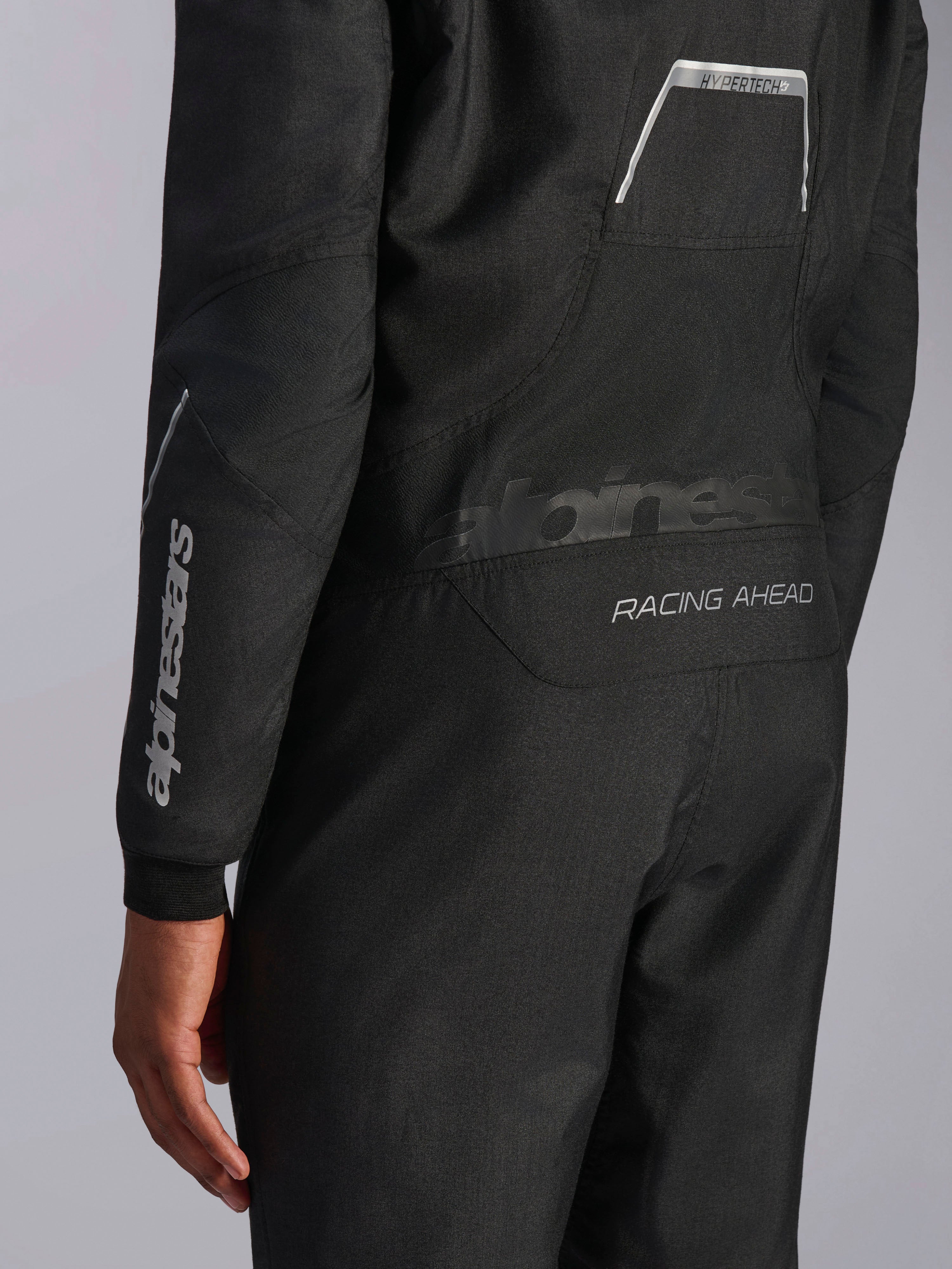 Zbliżenie na kombinezon Alpinestars Hypertech V3 w kolorze Black/Dark Shadow, pokazujące grafiki na plecach „RACING AHEAD” i „HYPERTECH V3”, białe logo na rękawie, profesjonalna odzież do wyścigów samochodowych