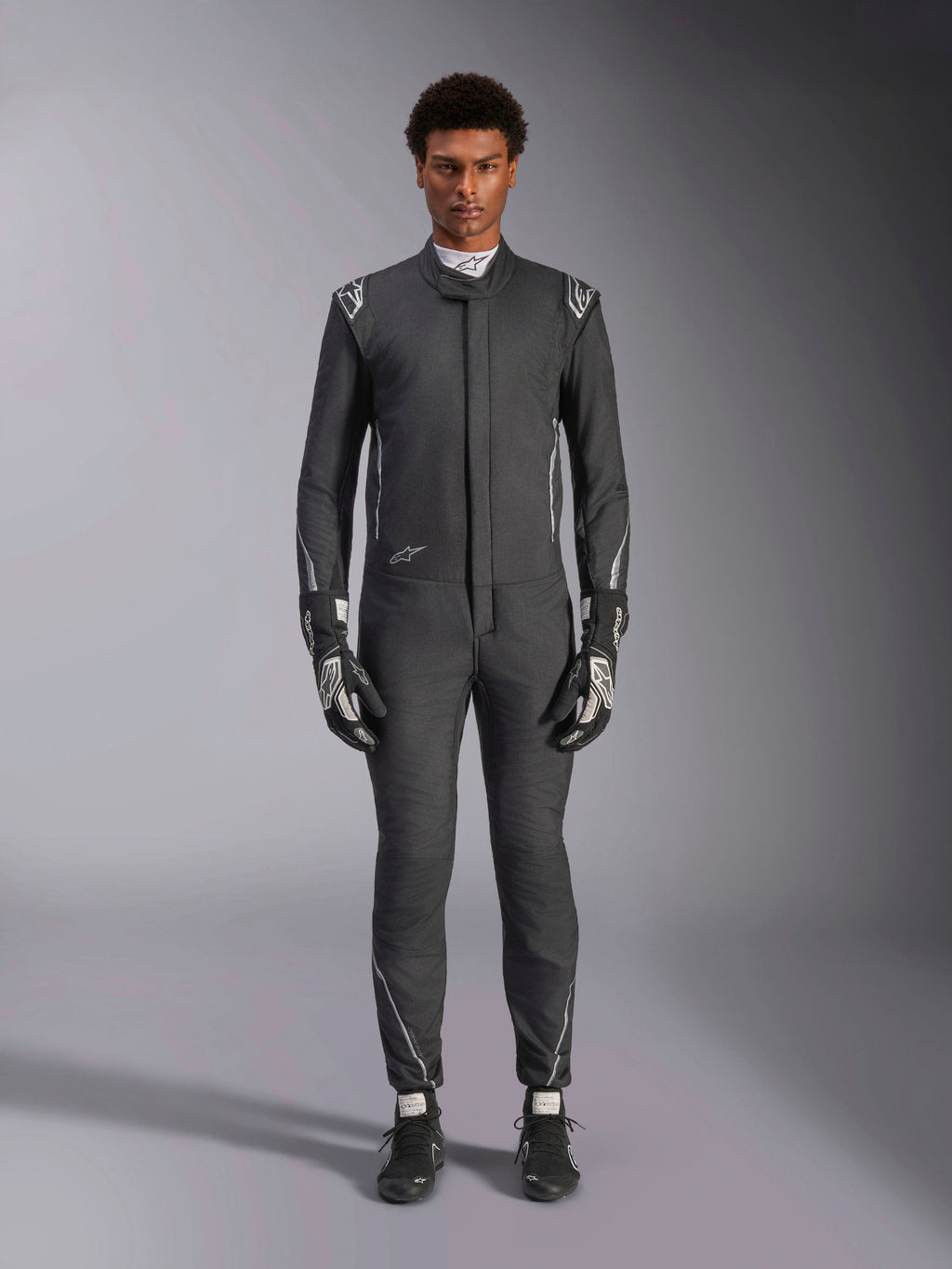 Alpinestars Hypertech V3 Suit, profesjonalny samochodowy kombinezon wyścigowy, czarny i Dark Shadow, prezentowany przez modela z dopasowanymi rękawicami i butami wyścigowymi, dopasowany sportowy krój ze srebrnymi akcentami i brandingiem, wyczynowa odzież motorsportowa
