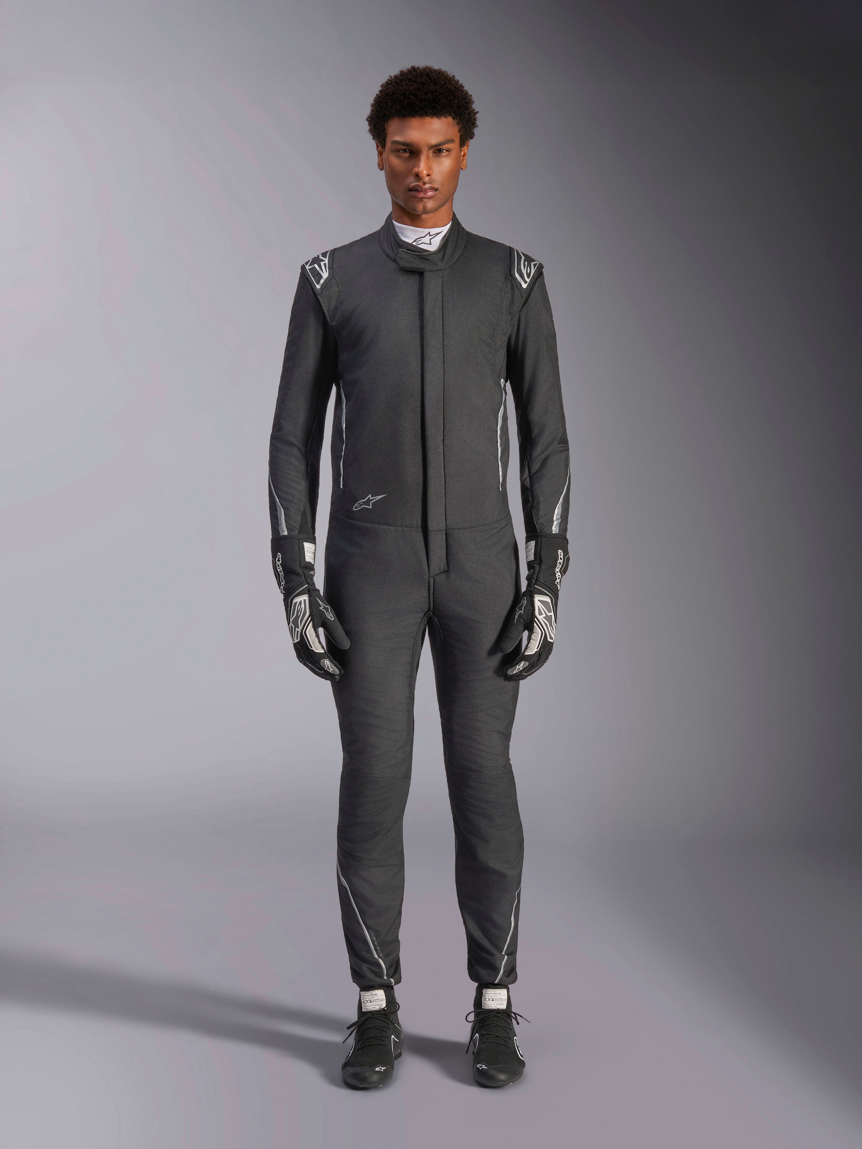 Alpinestars Hypertech V3 Suit, profesjonalny samochodowy kombinezon wyścigowy, czarny i Dark Shadow, prezentowany przez modela z dopasowanymi rękawicami i butami wyścigowymi, dopasowany sportowy krój ze srebrnymi akcentami i brandingiem, wyczynowa odzież motorsportowa