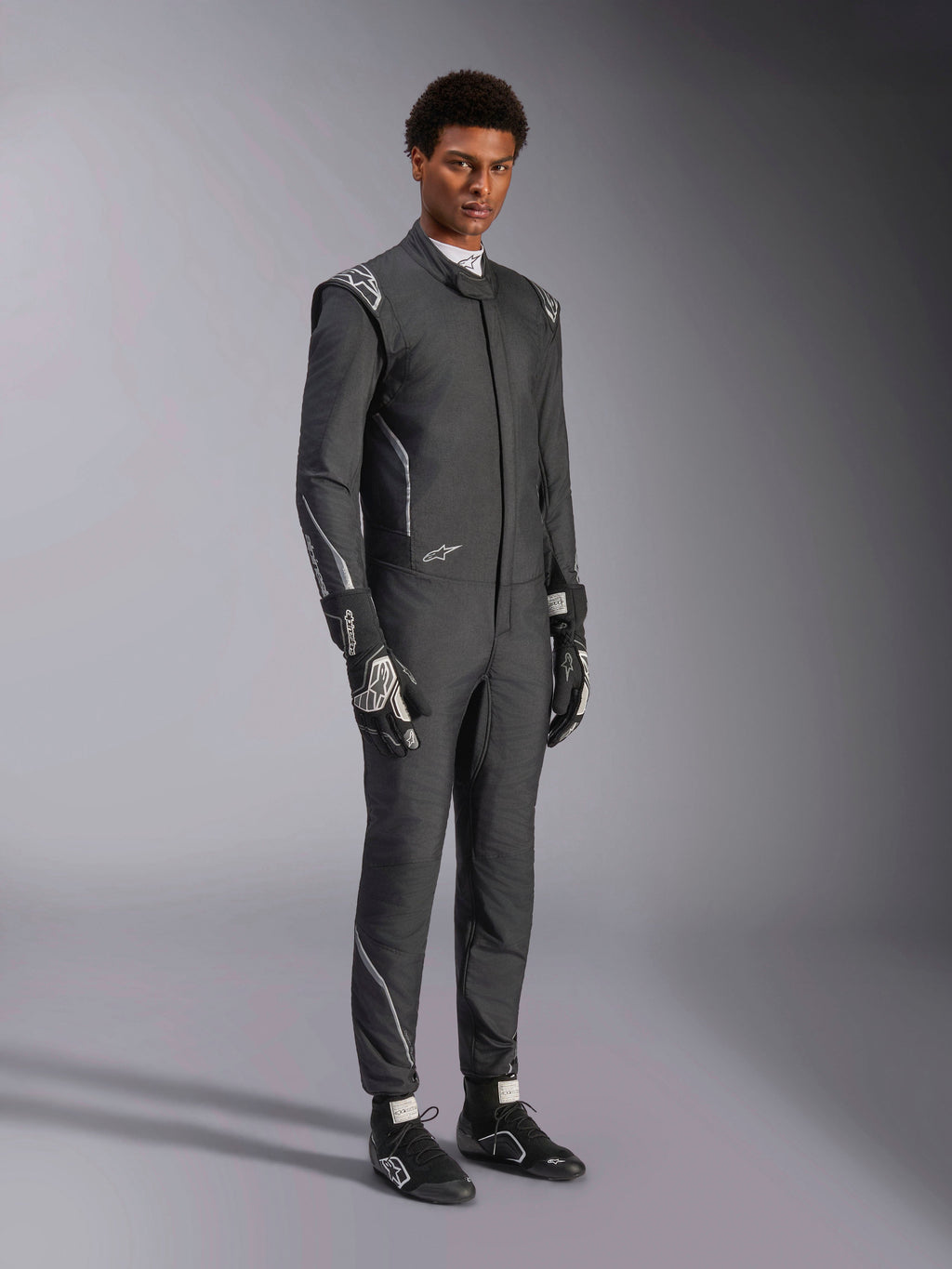 Kombinezon Alpinestars Hypertech V3, samochodowy kombinezon wyścigowy, kolor czarny i Dark Shadow, prezentowany przez modela z dopasowanymi czarnymi rękawicami i butami wyścigowymi, sportowy, dopasowany krój, widoczne logo Alpinestars i jasnoszare akcenty, profesjonalna odzież ochronna do sportów motorowych