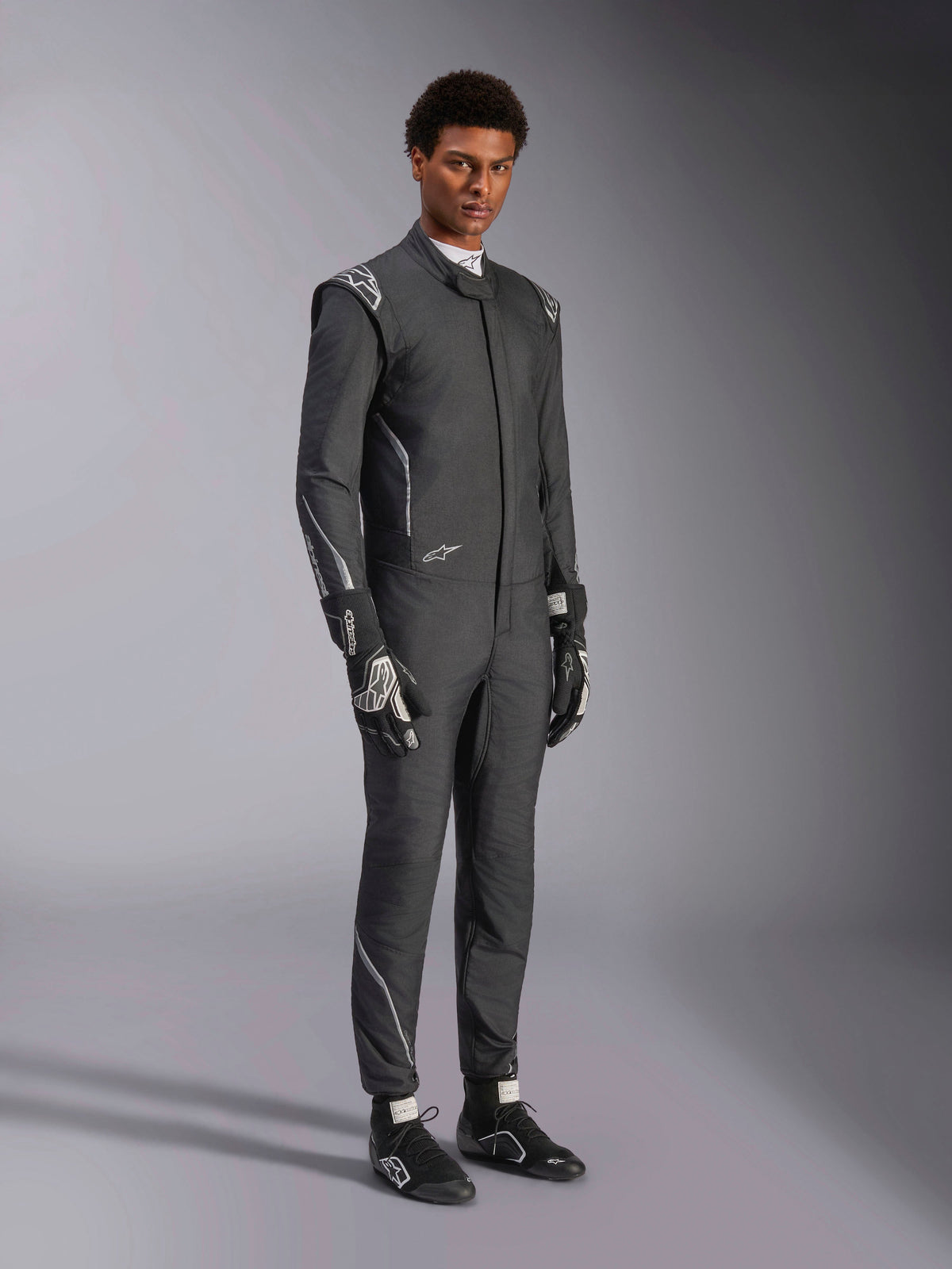 Kombinezon Alpinestars Hypertech V3, samochodowy kombinezon wyścigowy, kolor czarny i Dark Shadow, prezentowany przez modela z dopasowanymi czarnymi rękawicami i butami wyścigowymi, sportowy, dopasowany krój, widoczne logo Alpinestars i jasnoszare akcenty, profesjonalna odzież ochronna do sportów motorowych