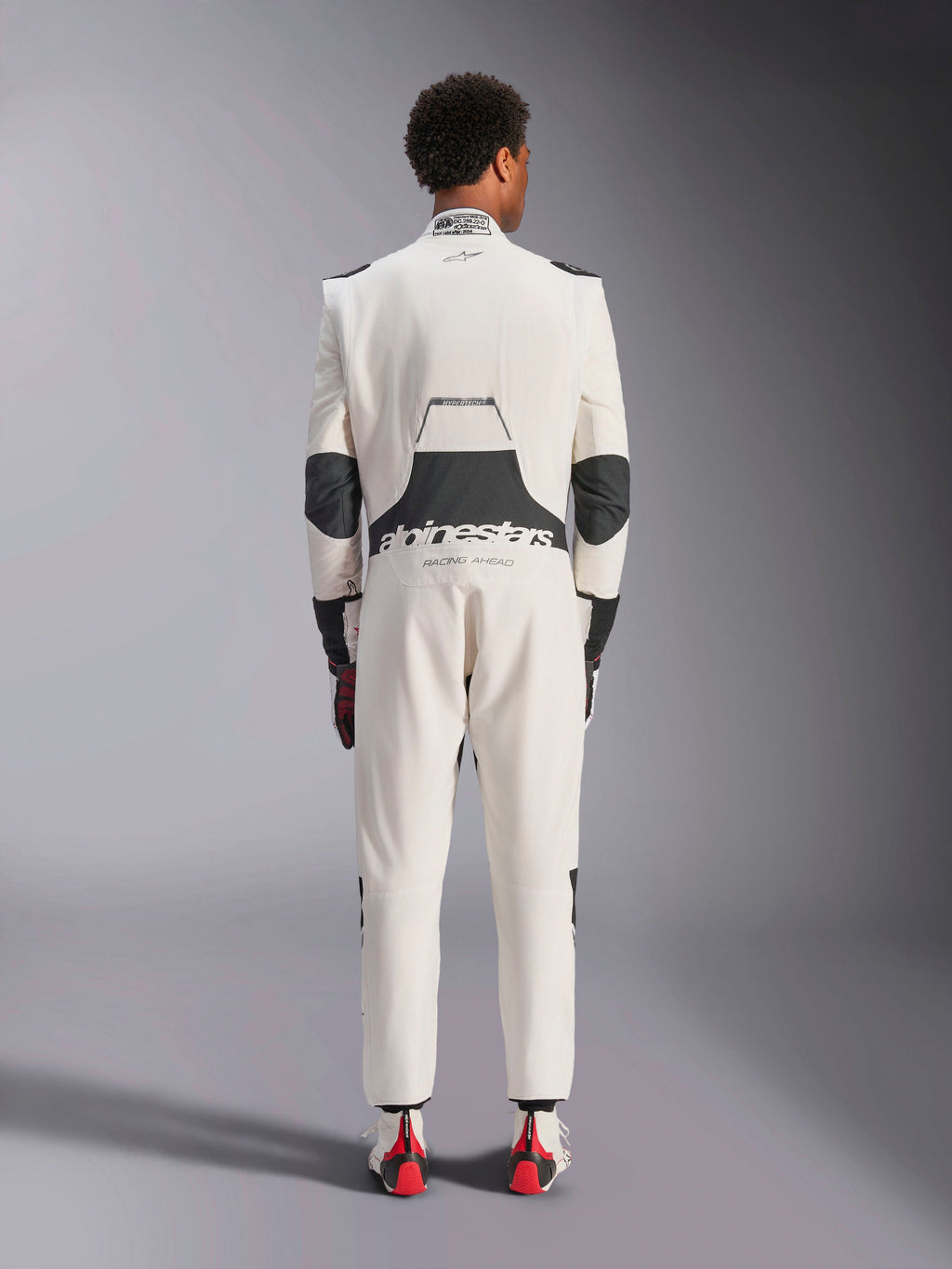 Widok z tyłu modela w kombinezonie Alpinestars Hypertech V3 w kolorze White Black Silver, prezentujący duży czarny elastyczny panel lędźwiowy z brandingiem „alpinestars” i „RACING AHEAD”, czarne wstawki na łokciach, etykietę certyfikacji FIA na kołnierzu oraz sportowy, dopasowany krój zaprojektowany do profesjonalnych wyścigów.