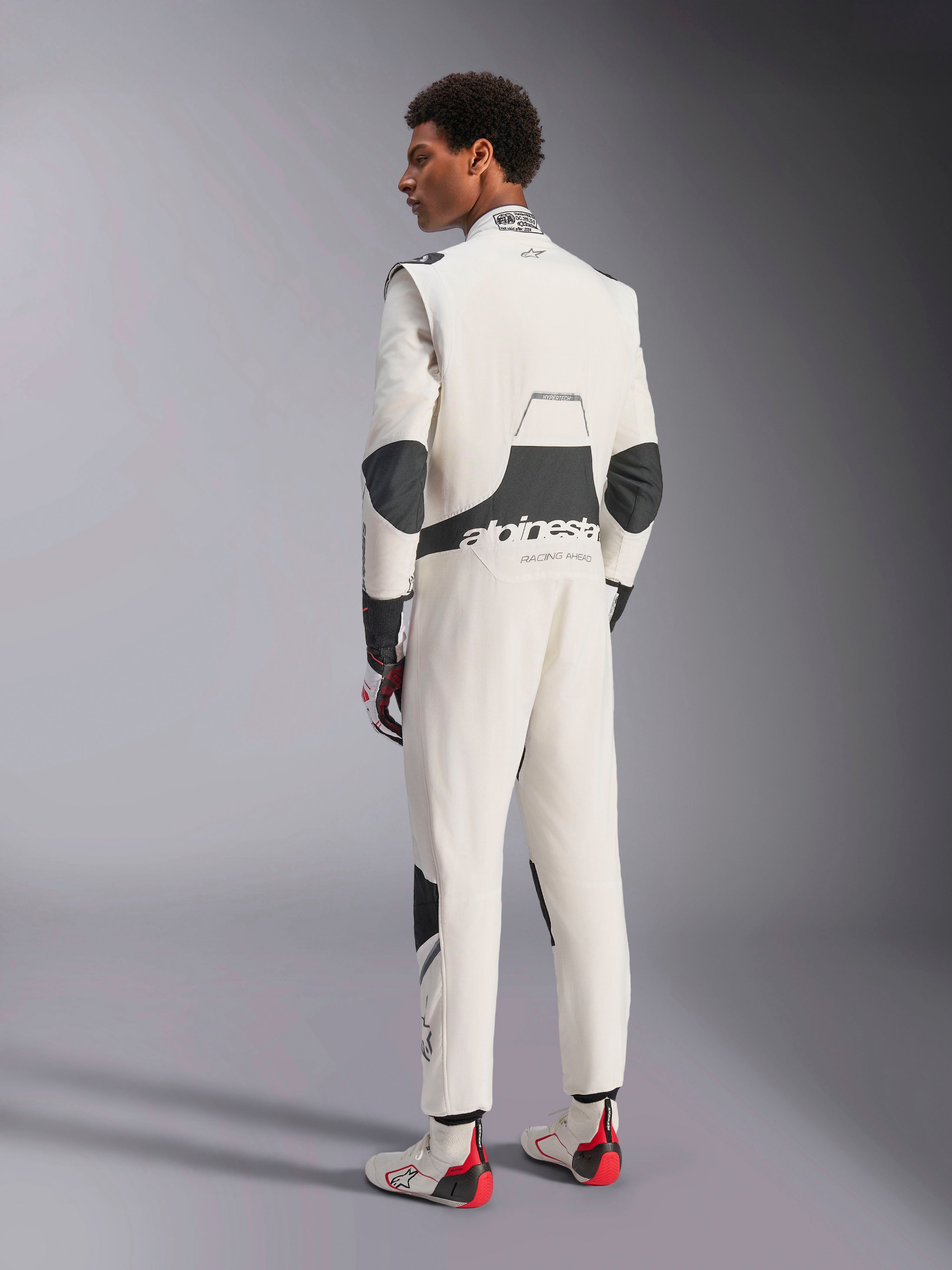 Kombinezon Alpinestars Hypertech V3, profesjonalny kombinezon do wyścigów samochodowych, biało-czarno-srebrny, widok modelu z tyłu ukazujący czarne panele elastyczne na lędźwiach i łokciach, branding Alpinestars na plecach, zestawiony z pasującymi rękawicami i butami wyścigowymi dla pełnego motorsportowego wyglądu