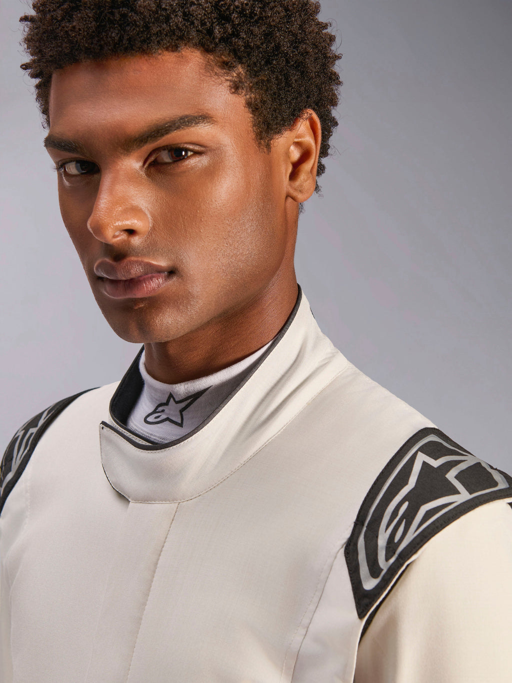 Zbliżenie na modela ubranego w kombinezon Alpinestars Hypertech V3 w kolorystyce White Black Silver, z wysokim kołnierzem z bezpiecznym zapięciem i logo Alpinestars na ramionach, profesjonalna odzież do wyścigów samochodowych.