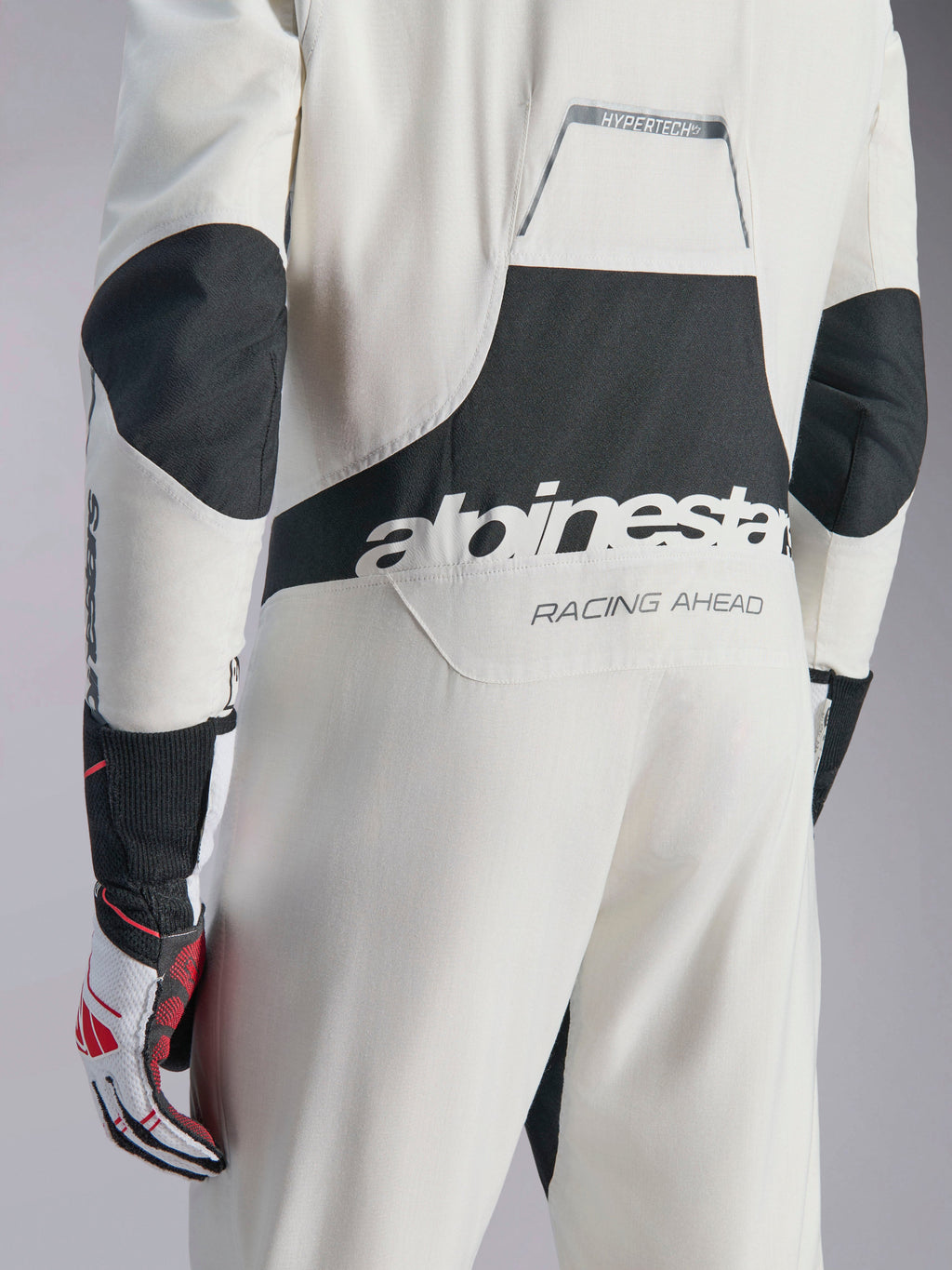 Kombinezon Alpinestars Hypertech V3, biało-czarno-srebrny kombinezon samochodowy, widok z tyłu z dużym czarnym logo „alpinestars” i napisem „RACING AHEAD”, czarne panele rozciągliwe na łokciach i w okolicy lędźwiowej, profesjonalna odzież ochronna do sportów motorowych