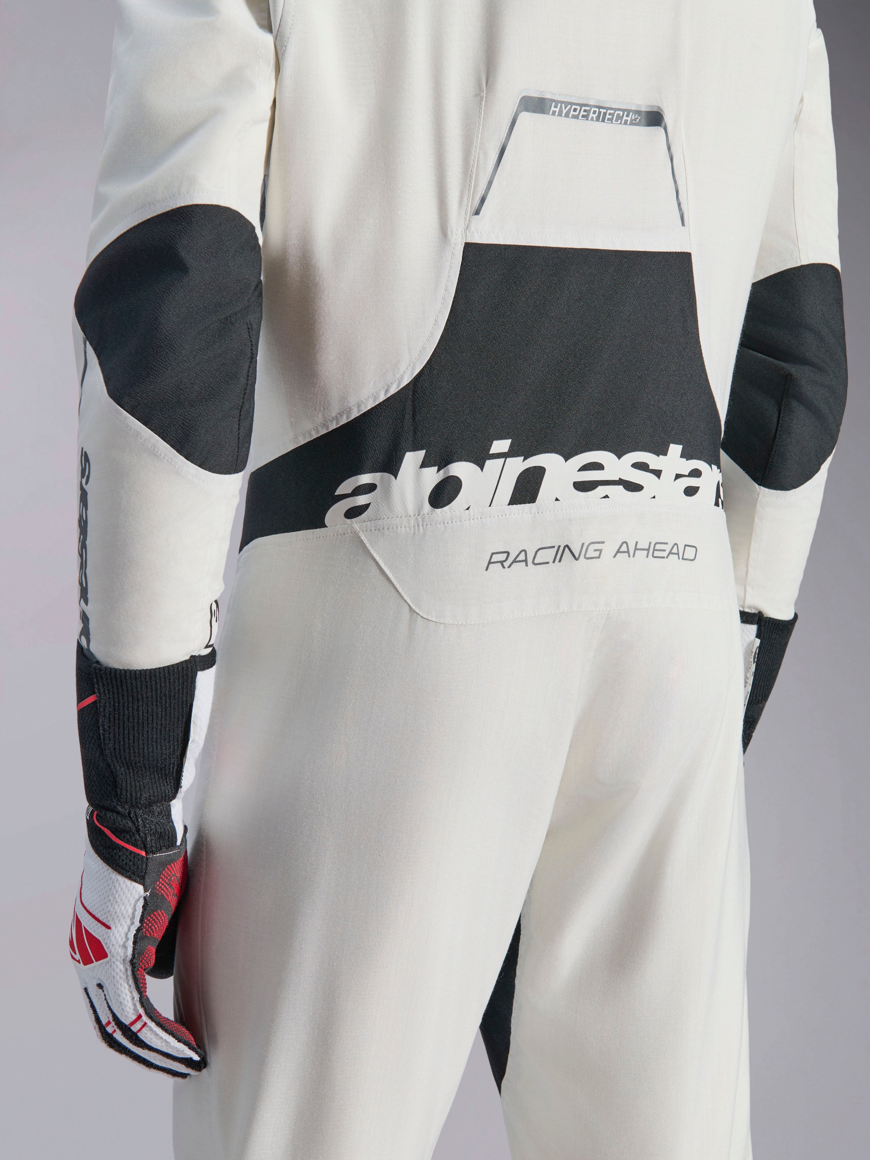 Kombinezon Alpinestars Hypertech V3, biało-czarno-srebrny kombinezon samochodowy, widok z tyłu z dużym czarnym logo „alpinestars” i napisem „RACING AHEAD”, czarne panele rozciągliwe na łokciach i w okolicy lędźwiowej, profesjonalna odzież ochronna do sportów motorowych