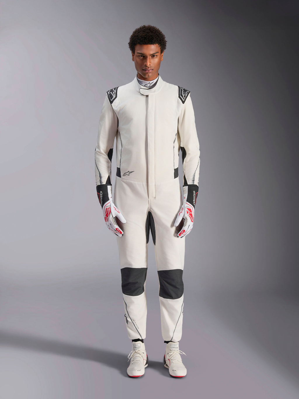 Kombinezon Alpinestars Hypertech V3, profesjonalny kombinezon samochodowy, biało-czarno-srebrny, prezentowany przez modela z czerwono-białymi rękawicami i pasującymi białymi butami wyścigowymi, z czarnymi kontrastowymi panelami i logo Alpinestars, sportowy, dopasowany krój