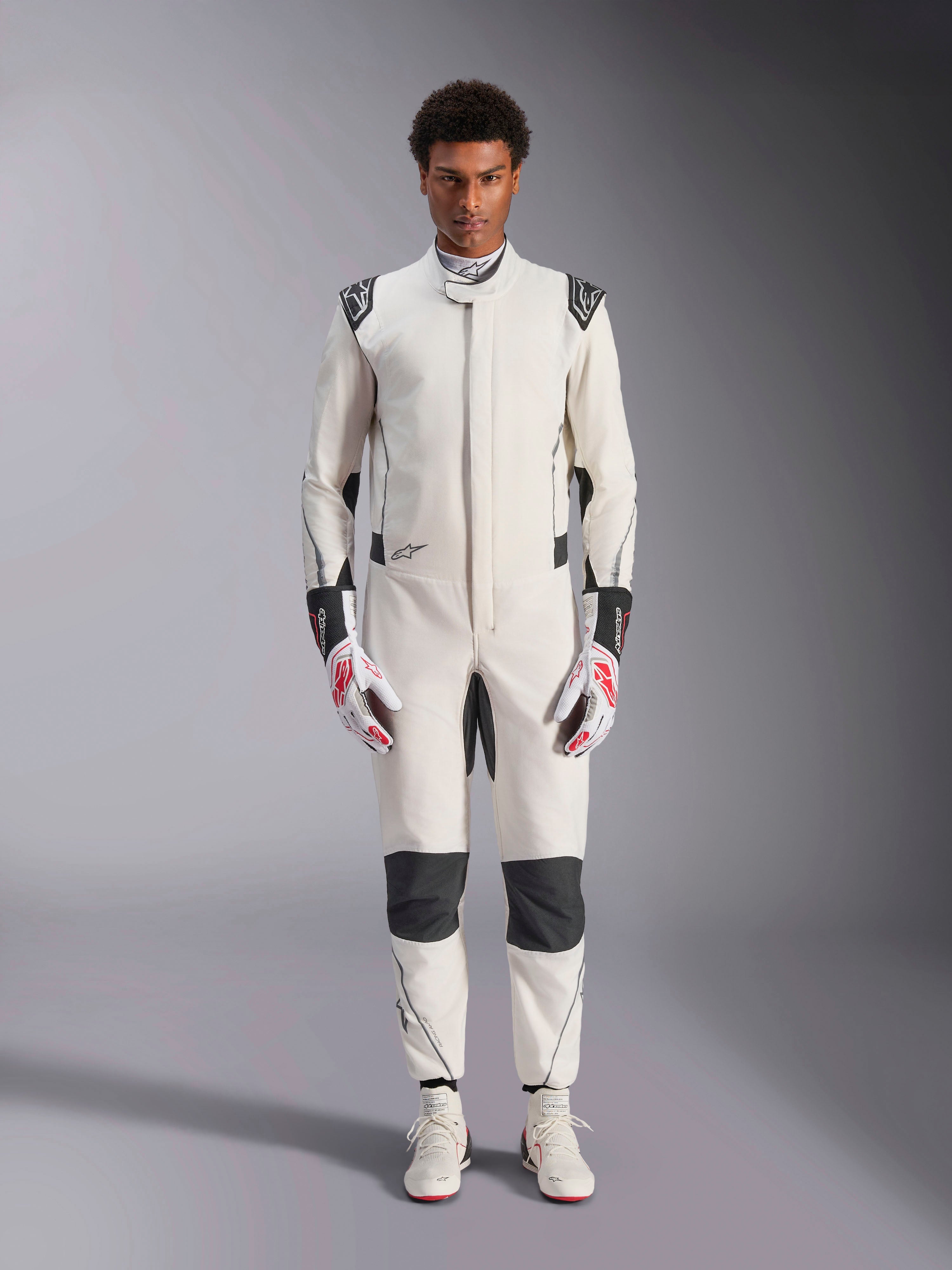 Kombinezon Alpinestars Hypertech V3, profesjonalny kombinezon samochodowy, biało-czarno-srebrny, prezentowany przez modela z czerwono-białymi rękawicami i pasującymi białymi butami wyścigowymi, z czarnymi kontrastowymi panelami i logo Alpinestars, sportowy, dopasowany krój