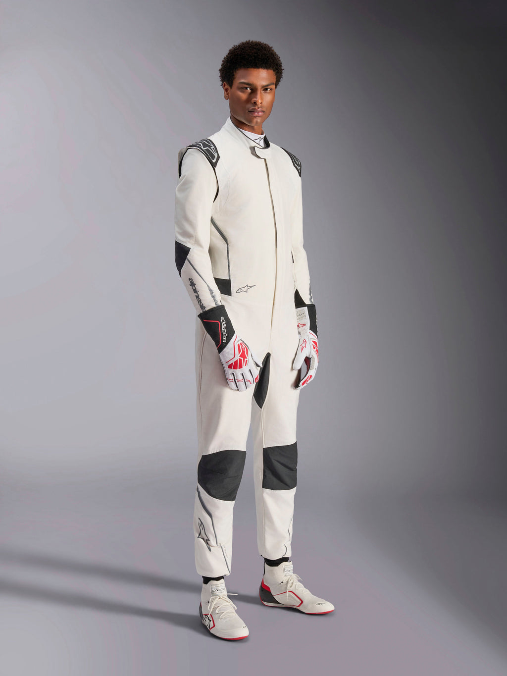 Męski model w kombinezonie Alpinestars Hypertech V3, biały z czarnymi i srebrnymi akcentami, profesjonalny strój do wyścigów samochodowych z czarnymi panelami na ramionach i kolanach, biało-czerwone rękawice wyścigowe i białe buty wyścigowe