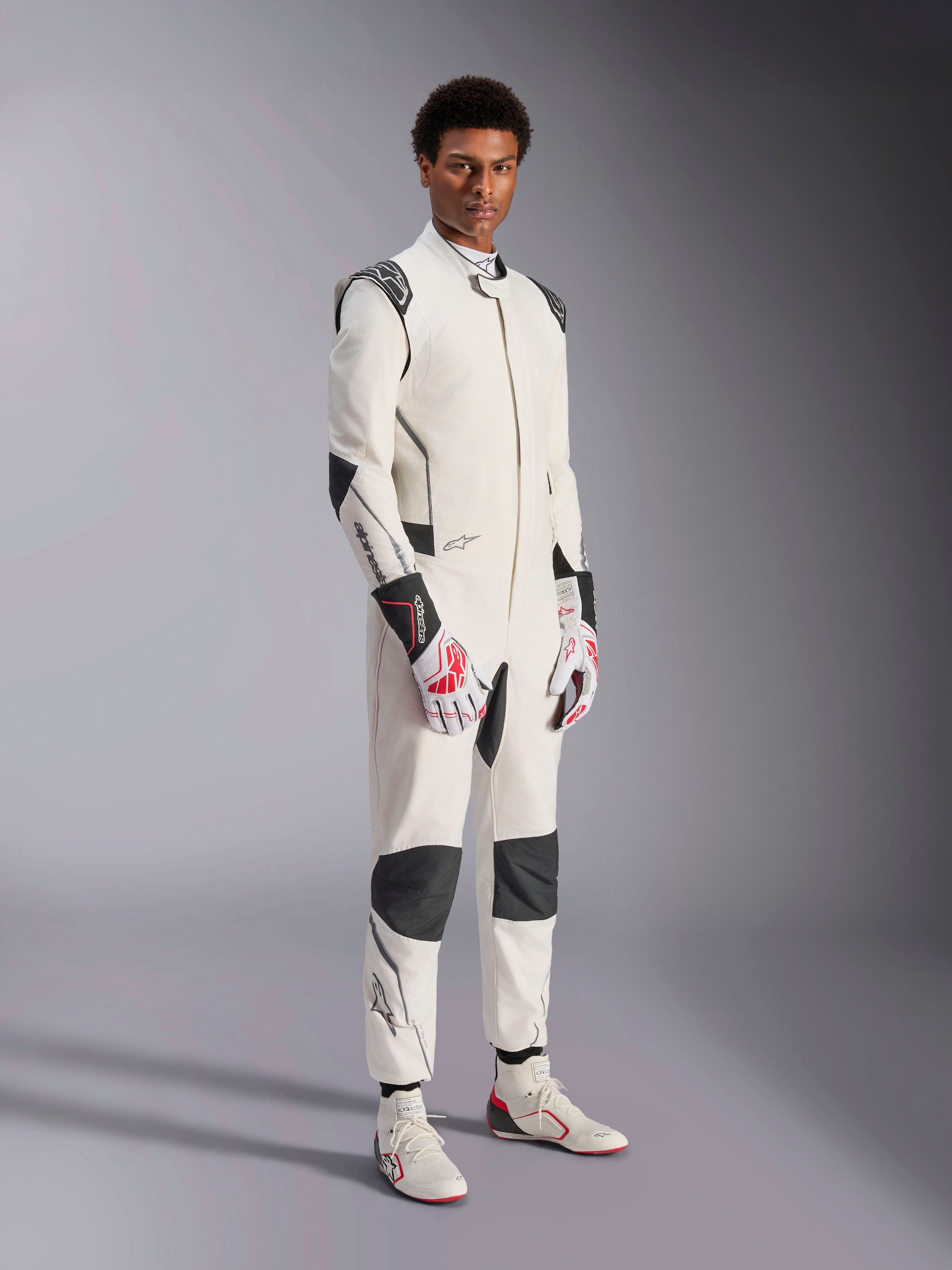 Męski model w kombinezonie Alpinestars Hypertech V3, biały z czarnymi i srebrnymi akcentami, profesjonalny strój do wyścigów samochodowych z czarnymi panelami na ramionach i kolanach, biało-czerwone rękawice wyścigowe i białe buty wyścigowe
