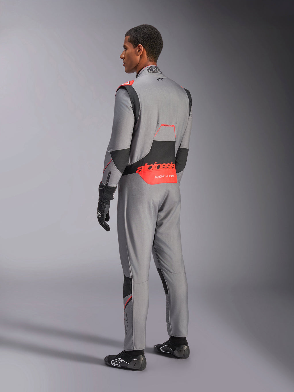 Kombinezon Alpinestars Hypertech V3 w kolorze Mid Gray Black Red Fluo, widok z tyłu pod kątem trzech czwartych, z wyraźnym czerwonym brandingiem Alpinestars w dolnej części pleców, czarnymi elastycznymi panelami i dopasowanym sportowym krojem do profesjonalnych wyścigów samochodowych.