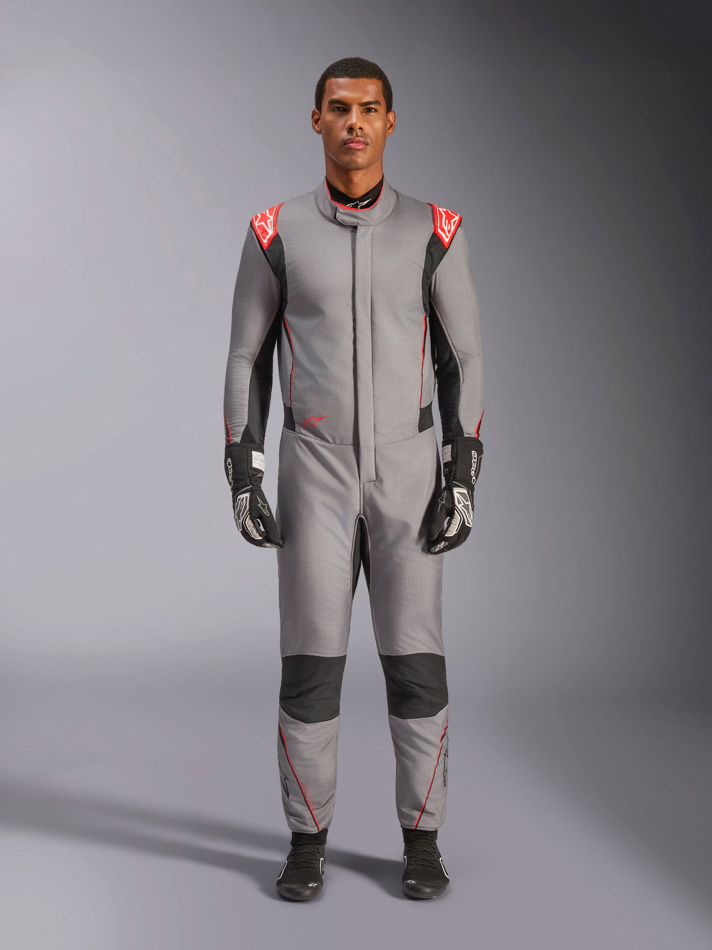 Alpinestars Hypertech V3 Suit, profesjonalny kombinezon wyścigowy w kolorze szarym, czarnym i jaskrawoczerwonym, prezentowany przez modela w czarnych rękawicach i butach wyścigowych, o sportowym, dopasowanym kroju, z jaskrawoczerwonymi akcentami na ramionach oraz czarnymi panelami na kolanach i po bokach.