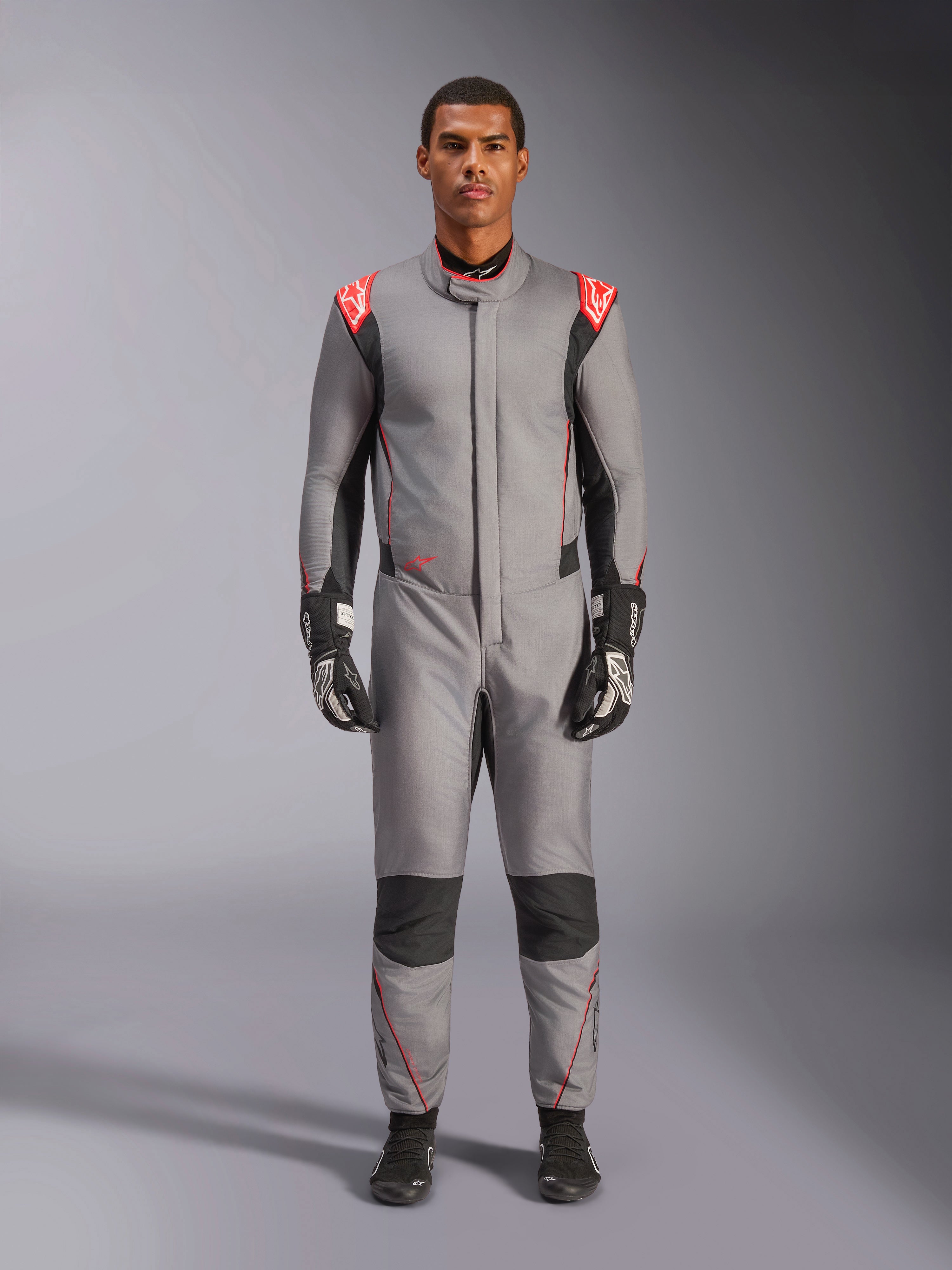 Alpinestars Hypertech V3 Suit, profesjonalny kombinezon wyścigowy w kolorze szarym, czarnym i jaskrawoczerwonym, prezentowany przez modela w czarnych rękawicach i butach wyścigowych, o sportowym, dopasowanym kroju, z jaskrawoczerwonymi akcentami na ramionach oraz czarnymi panelami na kolanach i po bokach.