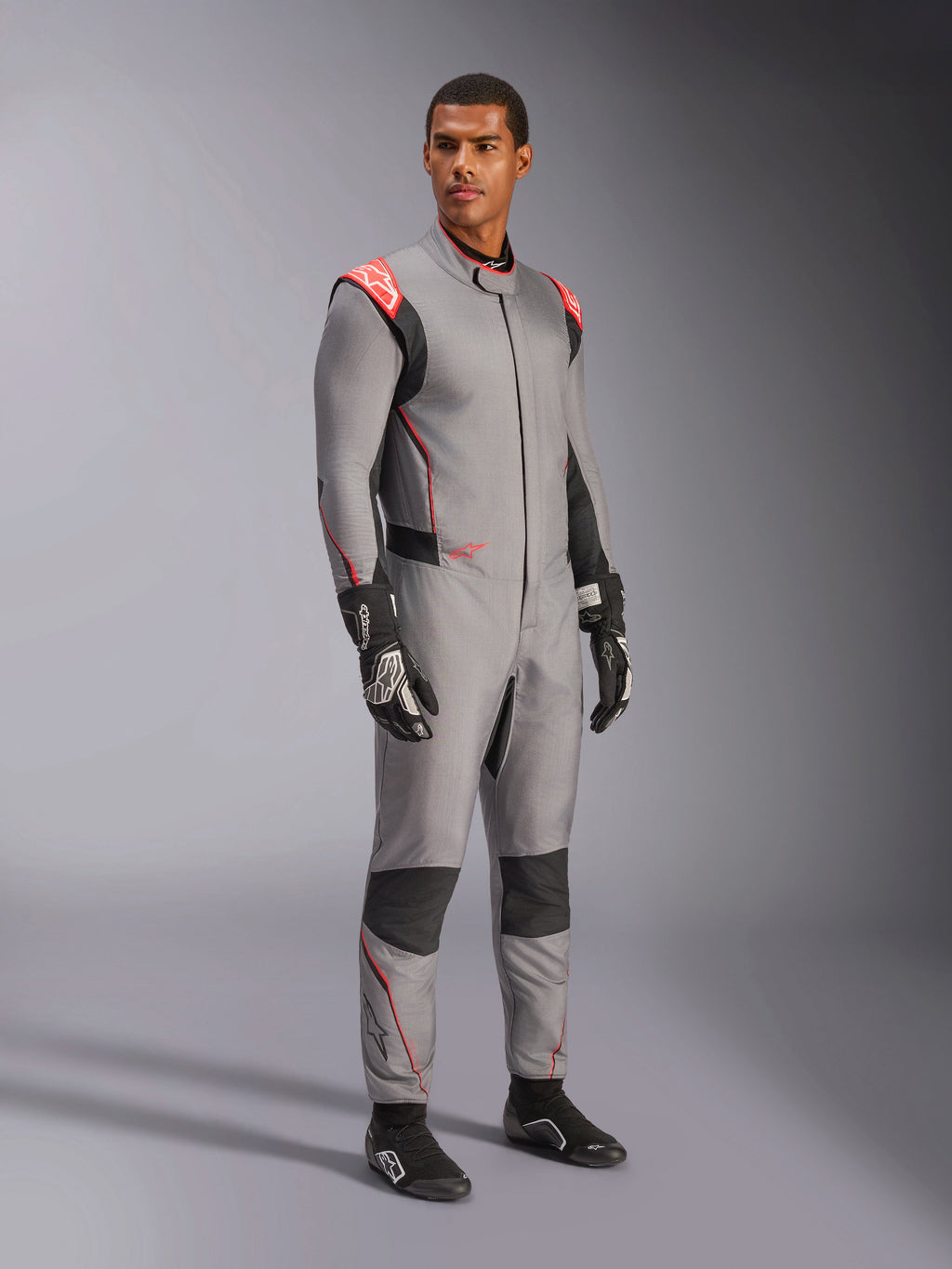 Męski model w kombinezonie Alpinestars Hypertech V3 w kolorze Mid Gray z czarnymi i czerwonymi akcentami Fluo, profesjonalny samochodowy kombinezon wyścigowy zestawiony z czarnymi rękawicami i butami wyścigowymi, charakteryzujący się lekką dwuwarstwową konstrukcją Nomex z czerwonymi detalami na ramionach i sportowym krojem