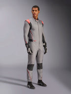 Męski model w kombinezonie Alpinestars Hypertech V3 w kolorze Mid Gray z czarnymi i czerwonymi akcentami Fluo, profesjonalny samochodowy kombinezon wyścigowy zestawiony z czarnymi rękawicami i butami wyścigowymi, charakteryzujący się lekką dwuwarstwową konstrukcją Nomex z czerwonymi detalami na ramionach i sportowym krojem