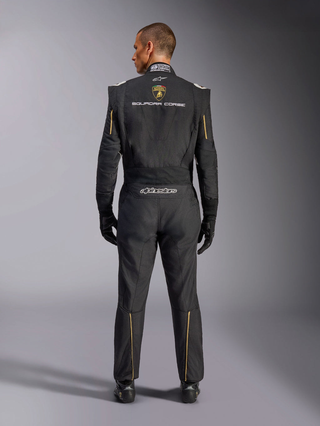 Kombinezon Alpinestars Limited Edition Lamborghini GP Tech V4 FIA, samochodowy kombinezon wyścigowy, czarny ze złotymi i srebrnymi akcentami, widok z tyłu prezentujący branding Lamborghini Squadra Corse, logo Alpinestars oraz złote lamówki na rękawach i nogawkach, profesjonalna odzież wyścigowa