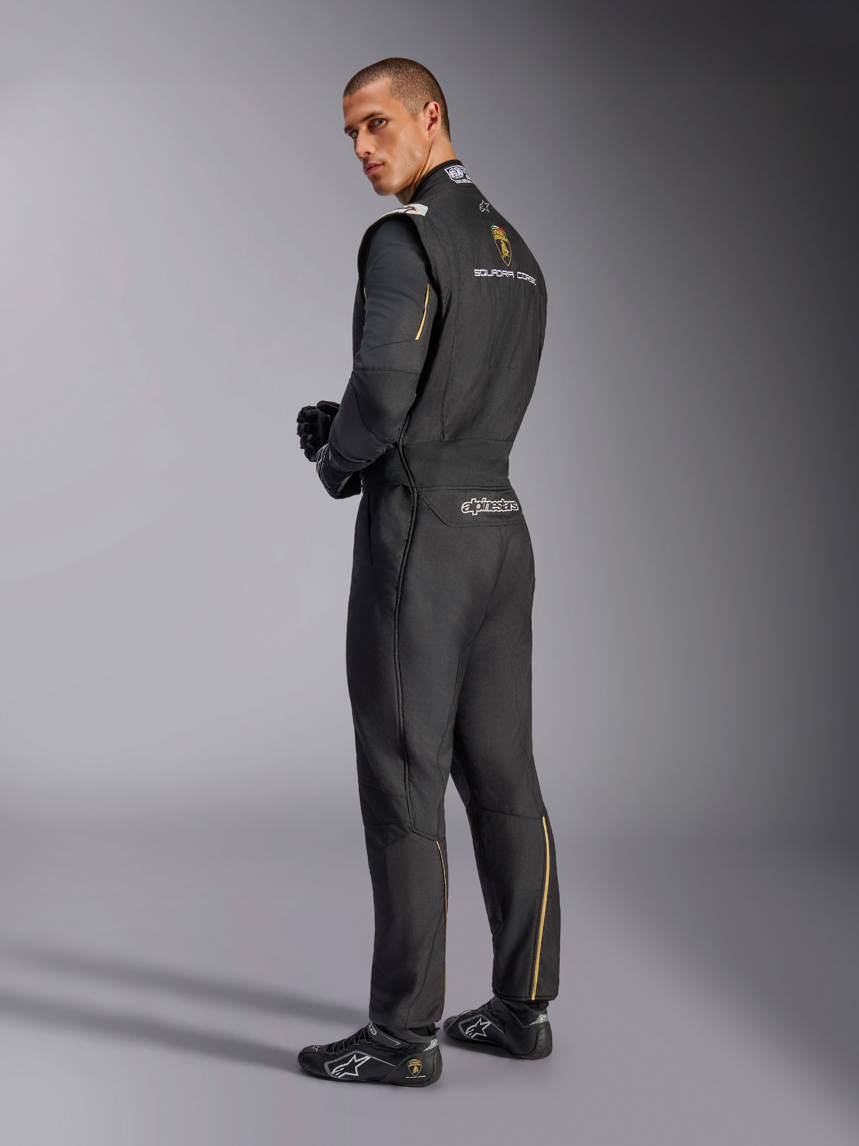 Kombinezon Alpinestars Limited Edition Lamborghini GP Tech V4, profesjonalny kombinezon wyścigowy, czarny ze srebrnymi i złotymi akcentami, widok z tyłu prezentujący branding Lamborghini Squadra Corse i Alpinestars, czarne rękawice i obuwie wyścigowe, smukły design motorsportowy