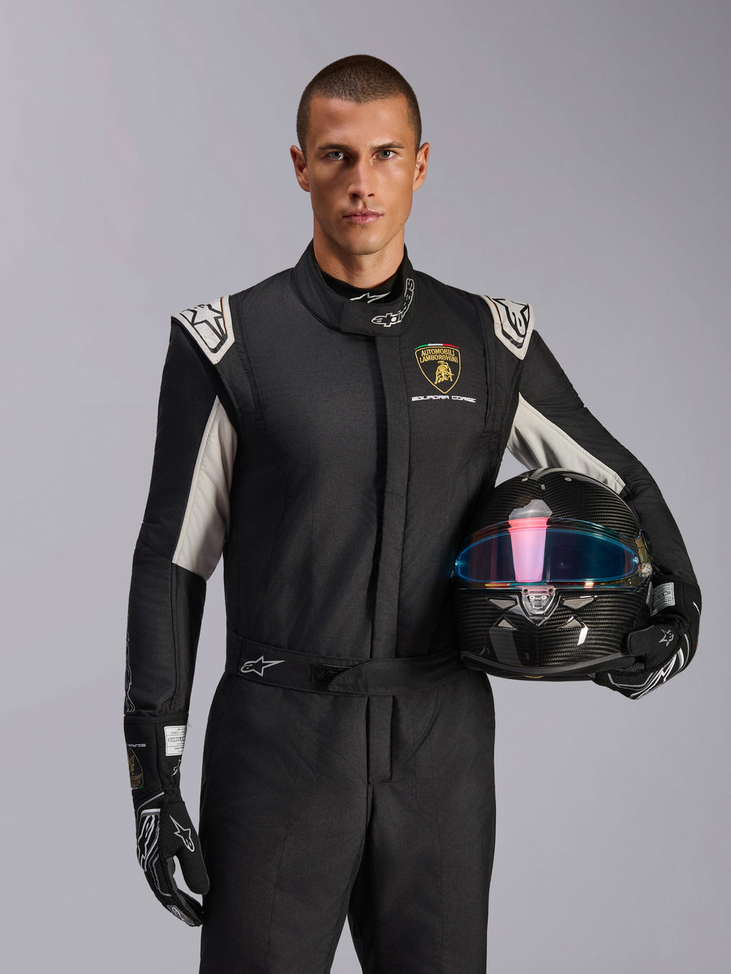 Model w kombinezonie Alpinestars Limited Edition Lamborghini GP Tech V4, czarny kombinezon wyścigowy ze srebrnymi akcentami i logo Lamborghini Squadra Corse, kask z włókna węglowego, czarne rękawice wyścigowe z białym logo, profesjonalny ubiór wyścigowy