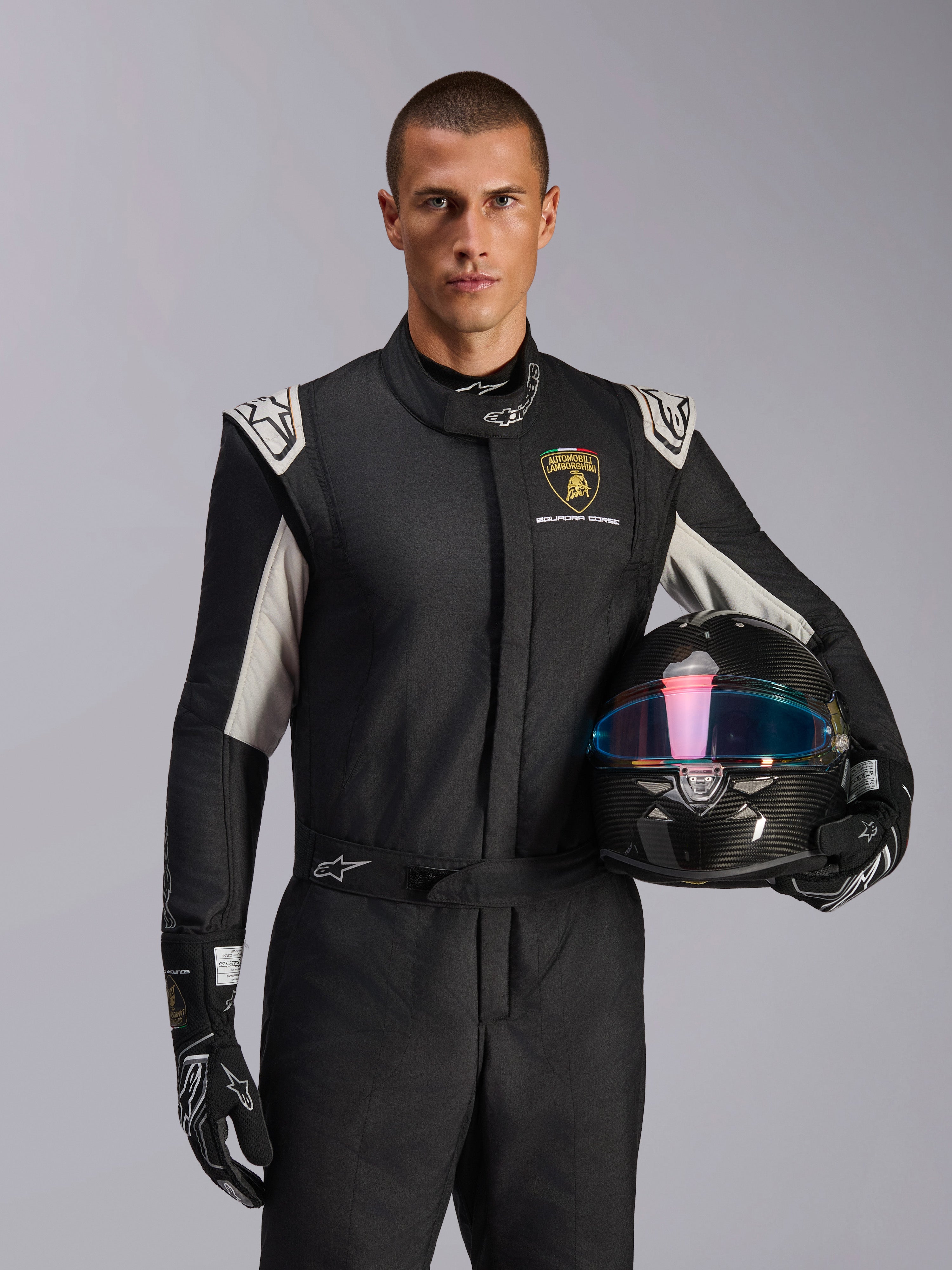 Model w kombinezonie Alpinestars Limited Edition Lamborghini GP Tech V4, czarny kombinezon wyścigowy ze srebrnymi akcentami i logo Lamborghini Squadra Corse, kask z włókna węglowego, czarne rękawice wyścigowe z białym logo, profesjonalny ubiór wyścigowy