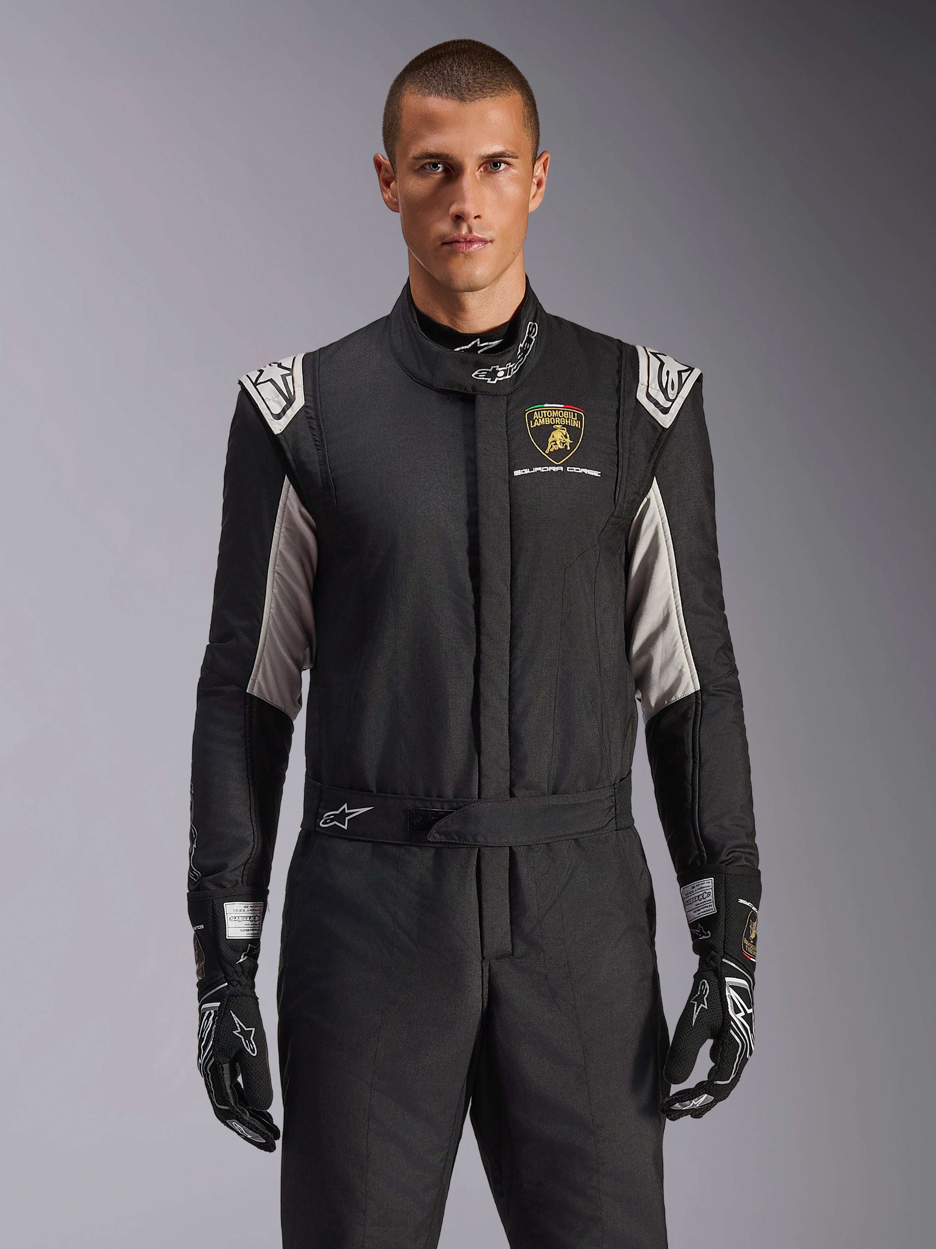 Alpinestars Limited Edition Lamborghini GP Tech V4 Suit, profesjonalny kombinezon wyścigowy w kolorze czarnym ze srebrnymi i złotymi akcentami, z tarczą Automobili Lamborghini Squadra Corse na piersi, noszony przez modela z dopasowanymi czarnymi rękawicami wyścigowymi