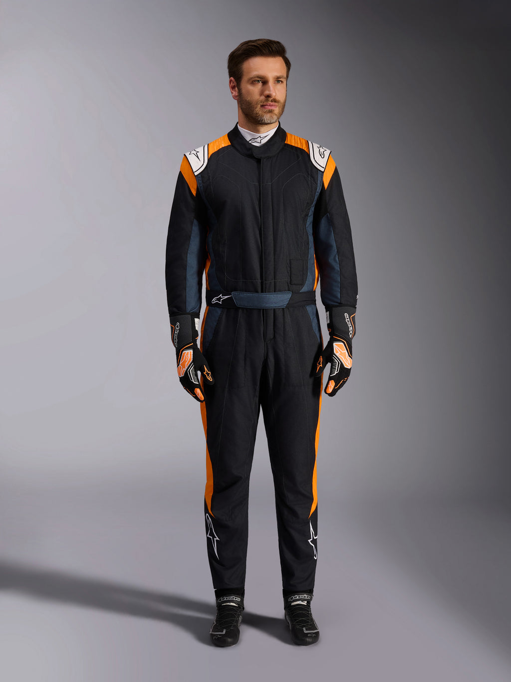 Alpinestars GP Pro Comp V2 FIA Suit, Black Asphalt and Orange Fluo auto racing suit, male model wearing full protective gear with matching racing gloves and shoes, features white Alpinestars logos and vibrant orange side panels, professional motorsport apparel