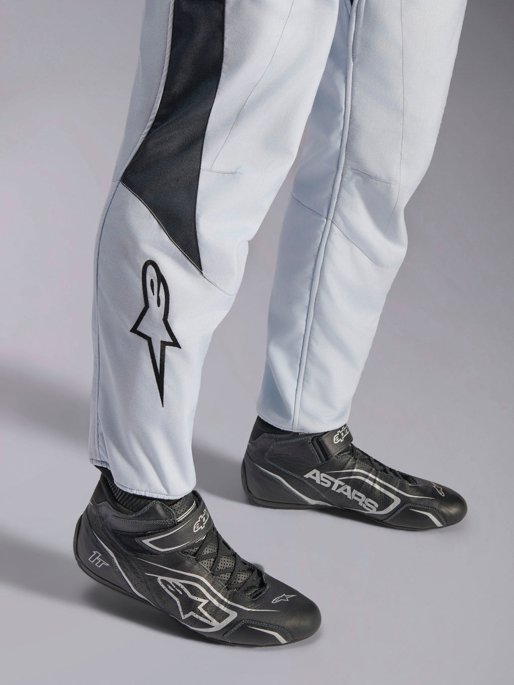 Alpinestars GP Pro Comp V2 FIA, kombinezon wyścigowy, szary i czarny, logo gwiazdy na nogawce i czarne buty wyścigowe.