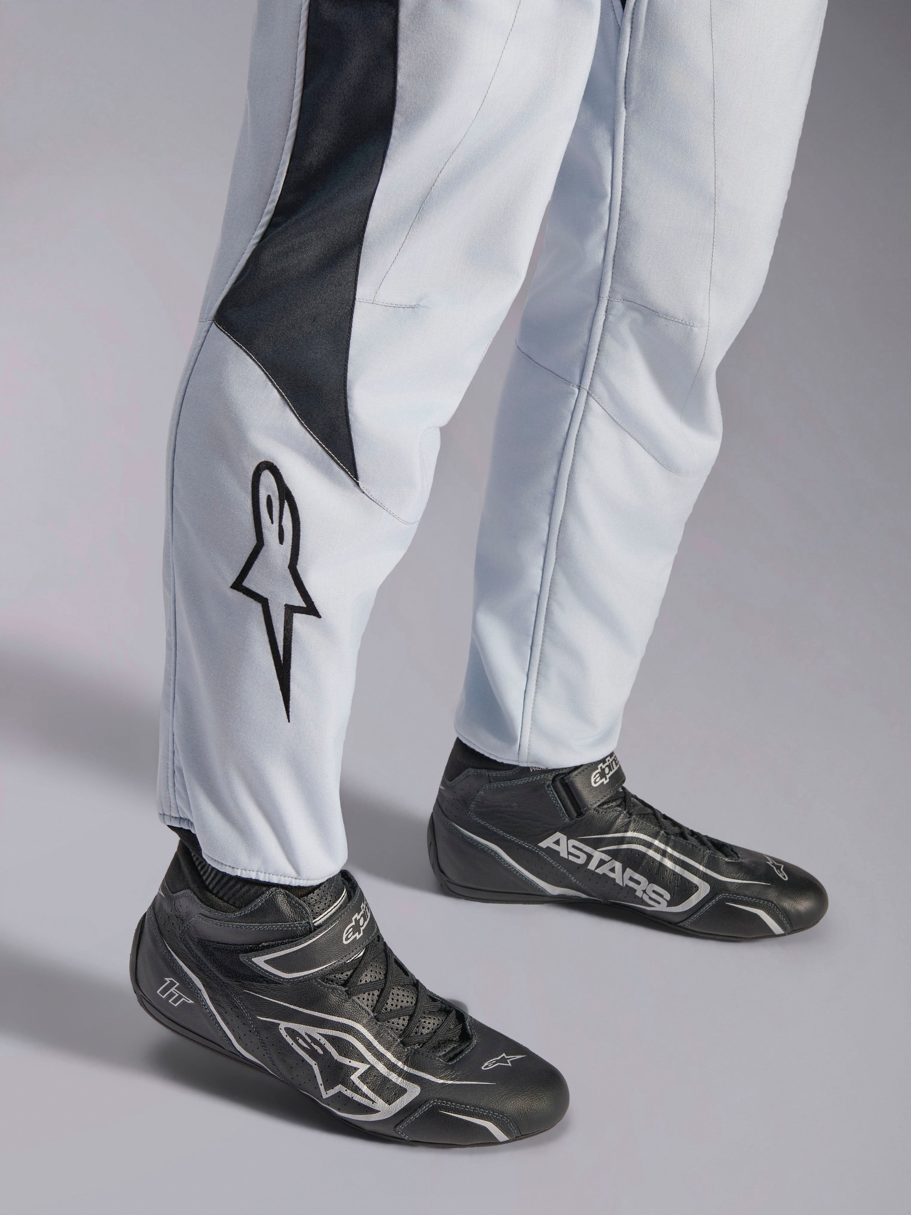 Alpinestars GP Pro Comp V2 FIA, kombinezon wyścigowy, szary i czarny, logo gwiazdy na nogawce i czarne buty wyścigowe.