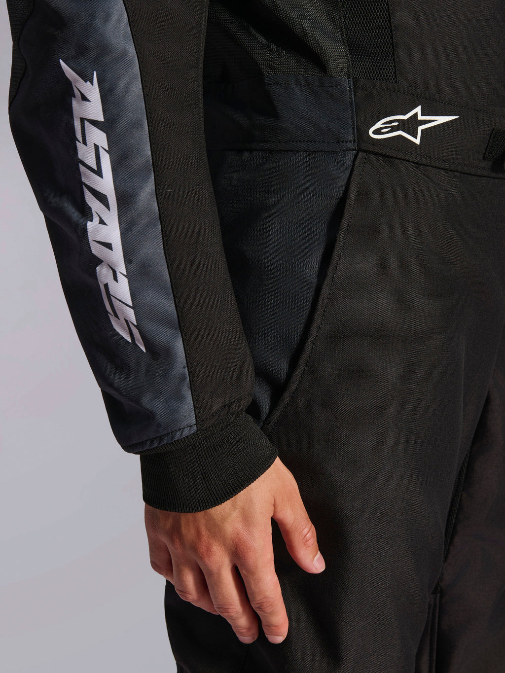 Kombinezon Alpinestars KMX-5 V3, kombinezon kartingowy, czarny i Dark Shadow, zbliżenie na biodro i ramię, biały napis ASTARS na rękawie, białe logo w pasie, z panelami z siatki dla wentylacji i dzianinowym mankietem