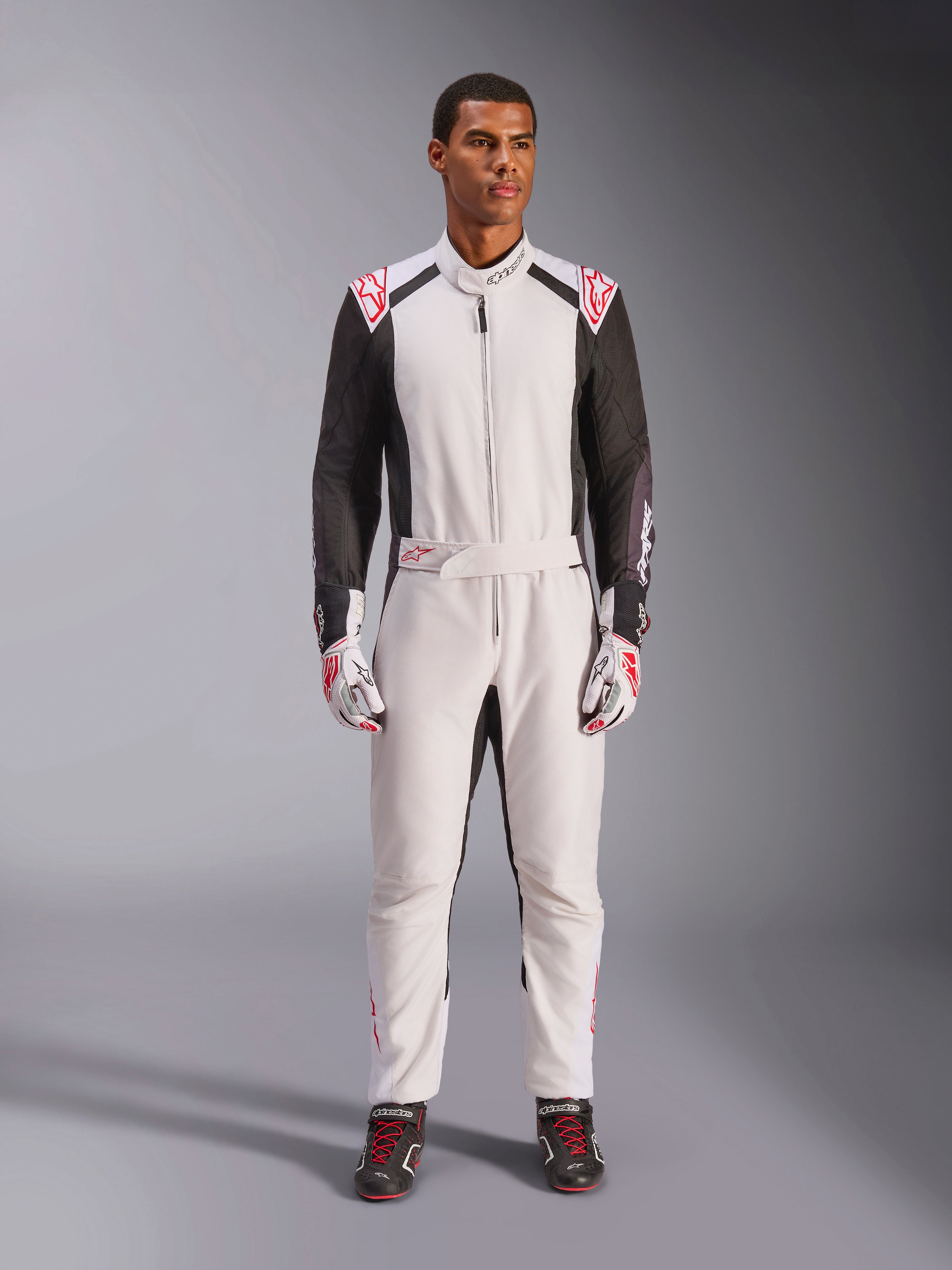 Kombinezon Alpinestars KMX-5 V3, jednoczęściowy kombinezon kartingowy, biało-czarno-czerwony, prezentowany przez modela wraz z dopasowanymi rękawicami i butami wyścigowymi, profesjonalny sprzęt motorsportowy do wyczynowej jazdy gokartami.
