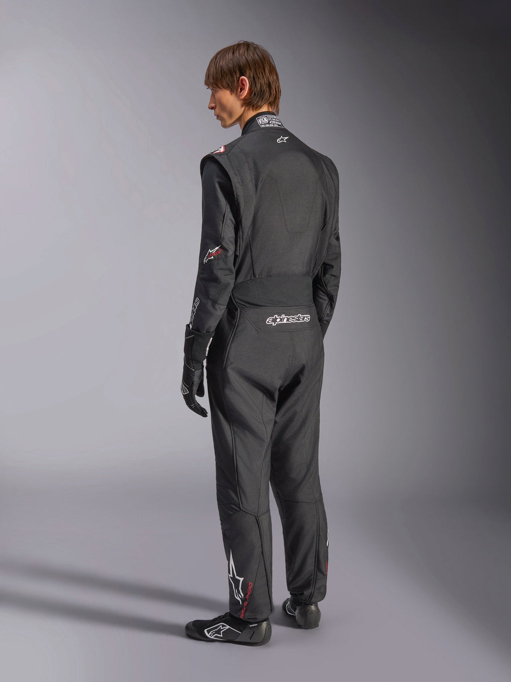 Kombinezon Alpinestars GP Tech V4, kombinezon samochodowy, czarny z białymi akcentami, widok z tyłu na modela ubranego w kombinezon, rękawice i buty wyścigowe, z białym logo na dole pleców i nogawce, profesjonalna odzież ochronna do sportów motorowych