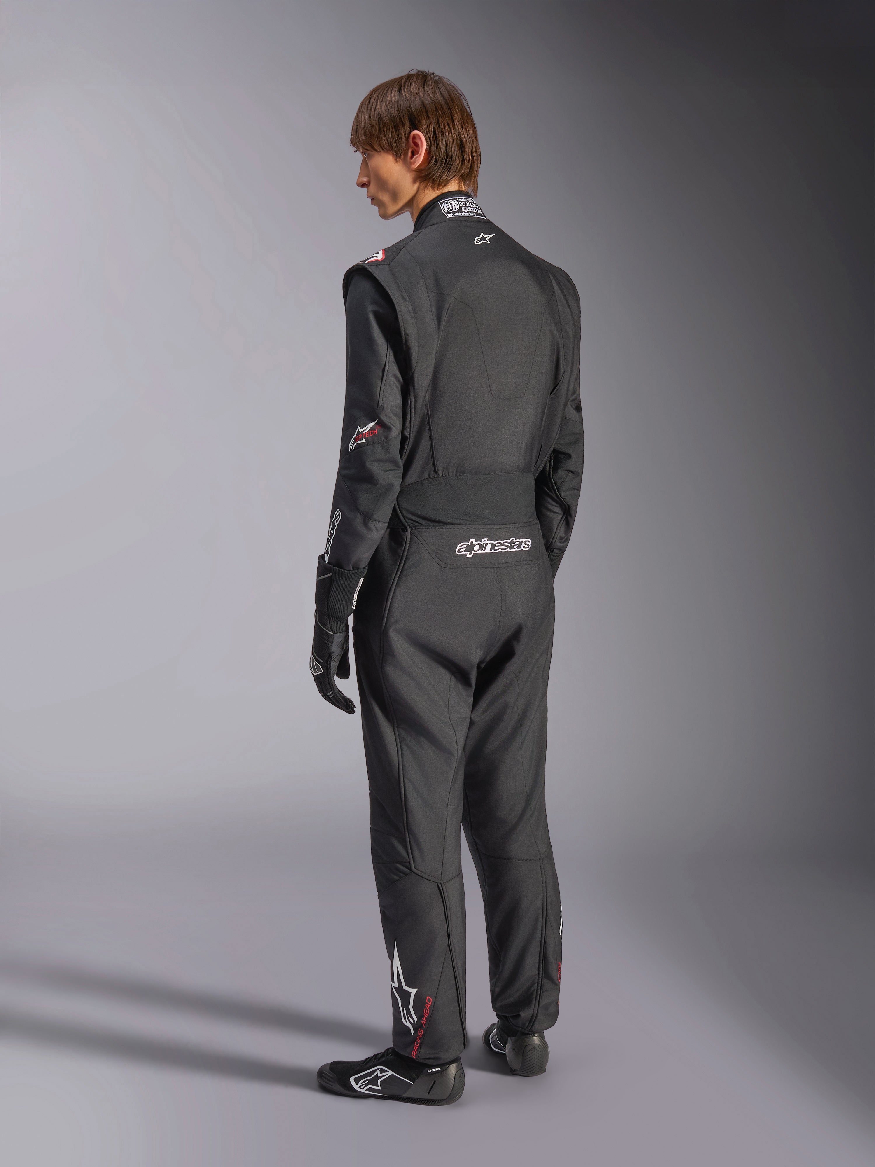 Kombinezon Alpinestars GP Tech V4, kombinezon samochodowy, czarny z białymi akcentami, widok z tyłu na modela ubranego w kombinezon, rękawice i buty wyścigowe, z białym logo na dole pleców i nogawce, profesjonalna odzież ochronna do sportów motorowych