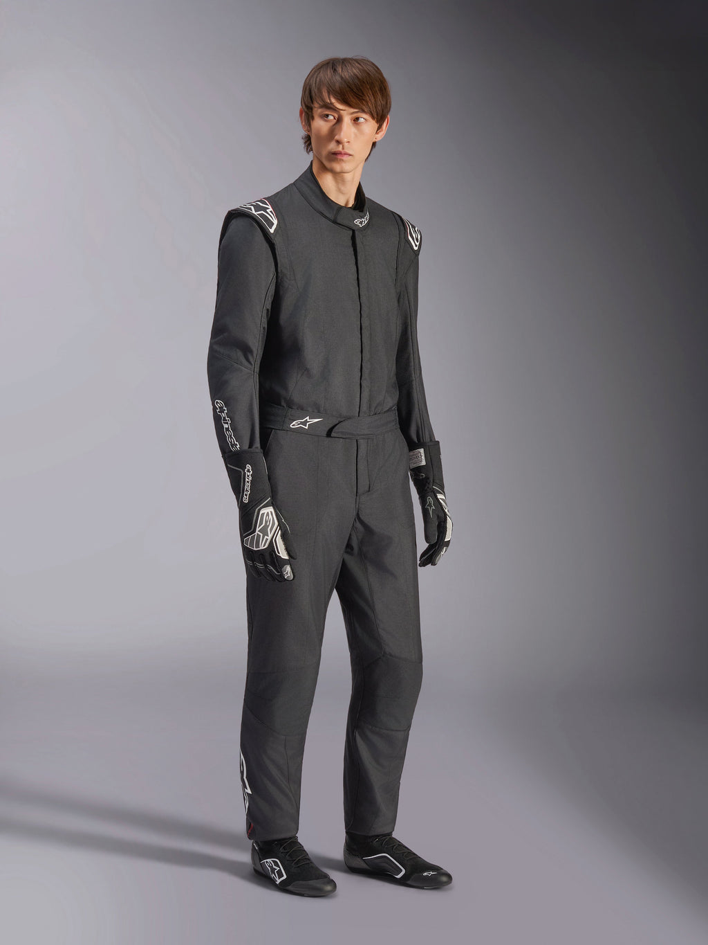 Alpinestars GP Tech V4 Suit, jednoczęściowy samochodowy kombinezon wyścigowy, czarno-biały, noszony z dopasowanymi rękawicami i obuwiem wyścigowym, białe logo na ramionach i przedramionach, profesjonalny design do sportów motorowych