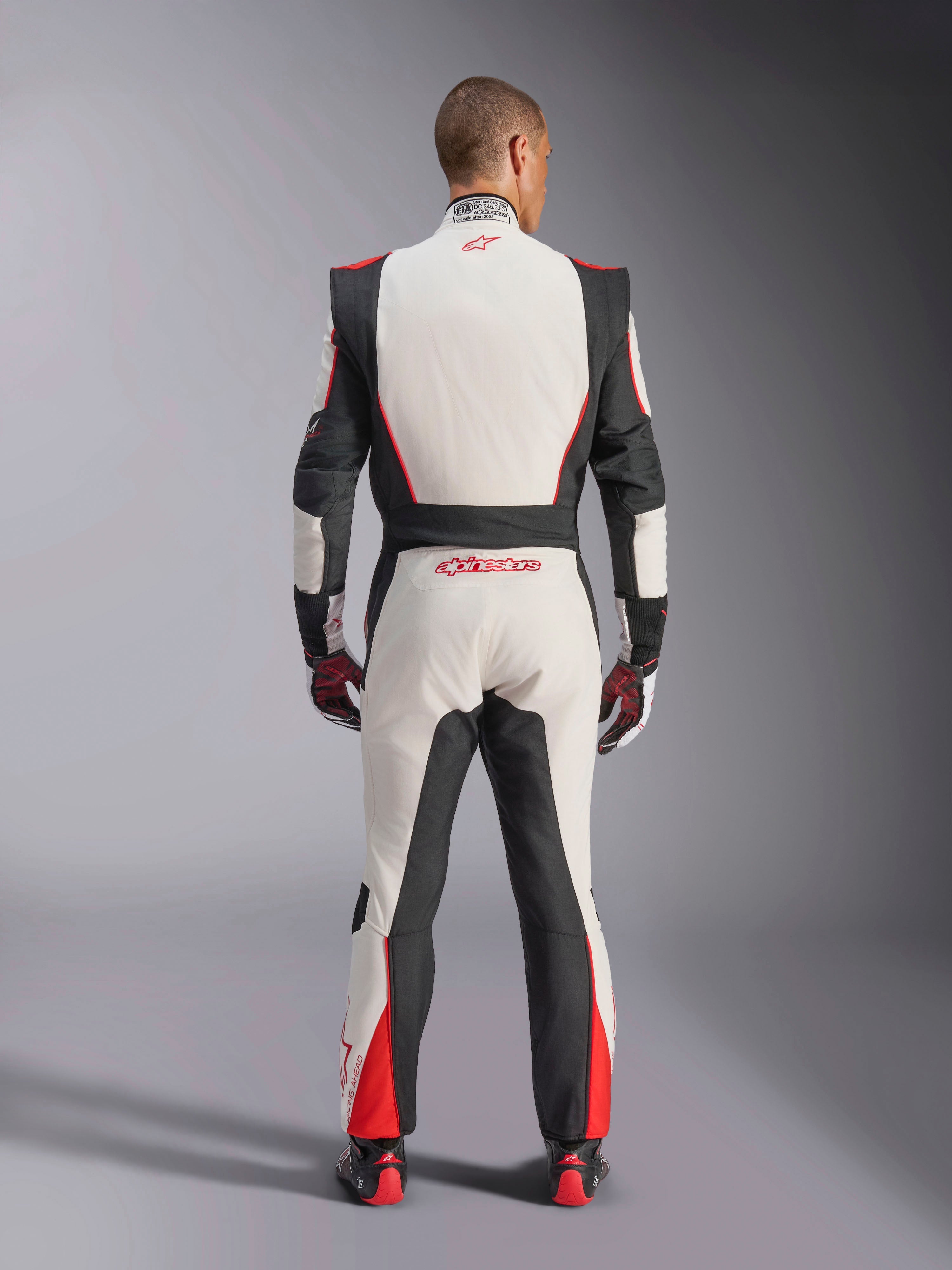 Alpinestars GP Tech V4 Suit, samochodowy kombinezon wyścigowy, biało-czarno-czerwony, widok od tyłu na kierowcę w jednoczęściowym kombinezonie, czarne elastyczne panele po bokach i na ramionach, czerwone akcenty na dolnej części nogawek, branding Alpinestars na dole pleców i kołnierzu, profesjonalny sprzęt wyścigowy z certyfikatem FIA