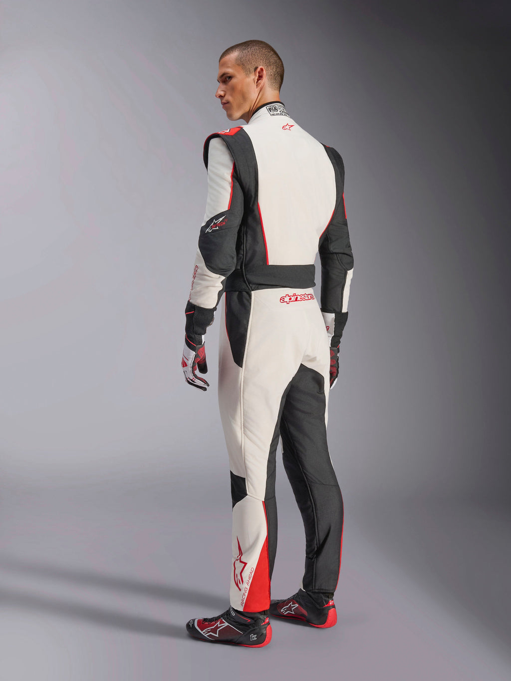 Kombinezon Alpinestars GP Tech V4, biało-czarno-czerwony kombinezon wyścigowy, widok z tyłu pokazujący ergonomiczne panele elastyczne na dolnej części pleców i nogawkach, w zestawie z dopasowanymi rękawicami i butami wyścigowymi, profesjonalna odzież motorsportowa.