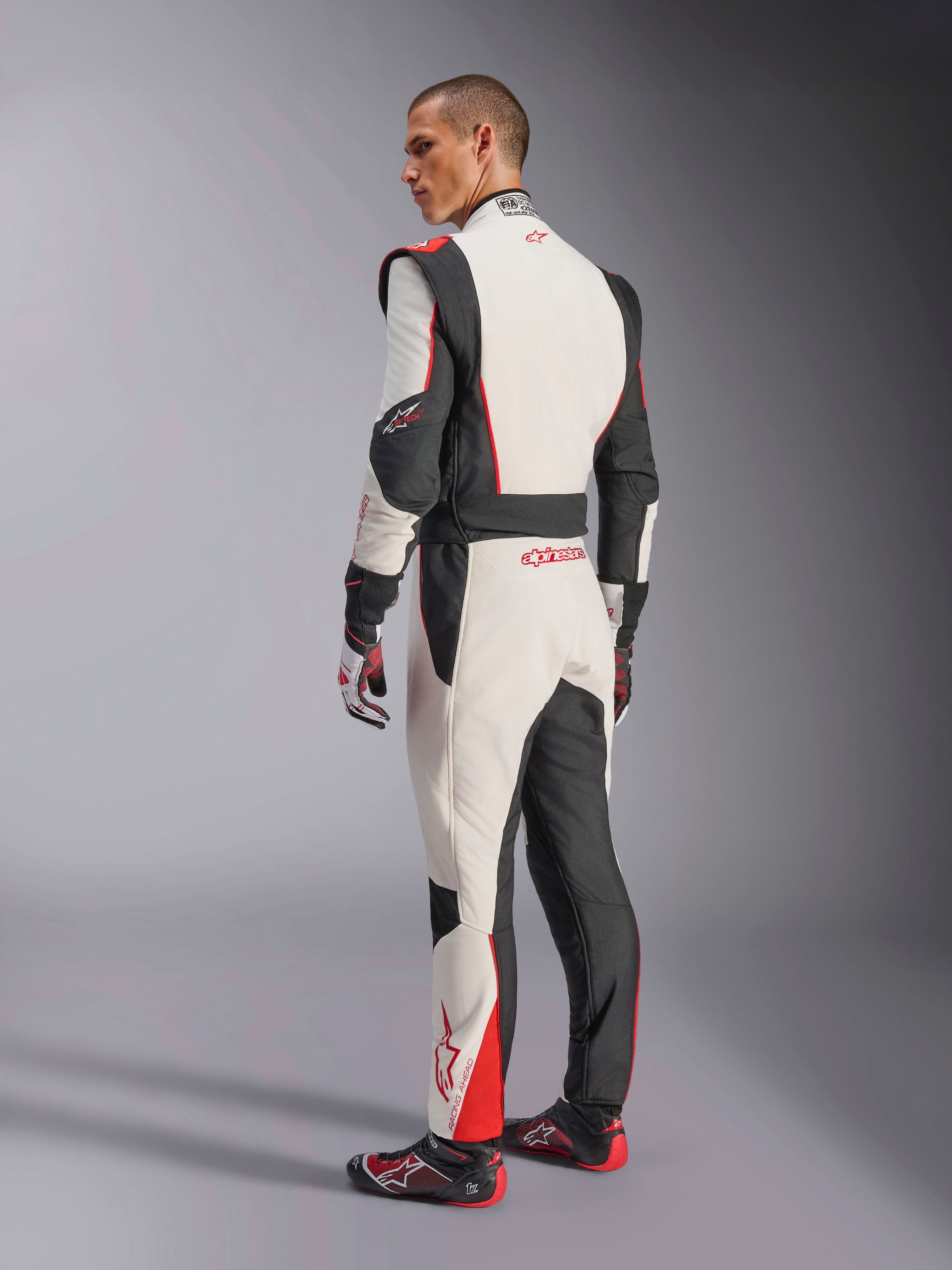 Kombinezon Alpinestars GP Tech V4, biało-czarno-czerwony kombinezon wyścigowy, widok z tyłu pokazujący ergonomiczne panele elastyczne na dolnej części pleców i nogawkach, w zestawie z dopasowanymi rękawicami i butami wyścigowymi, profesjonalna odzież motorsportowa.
