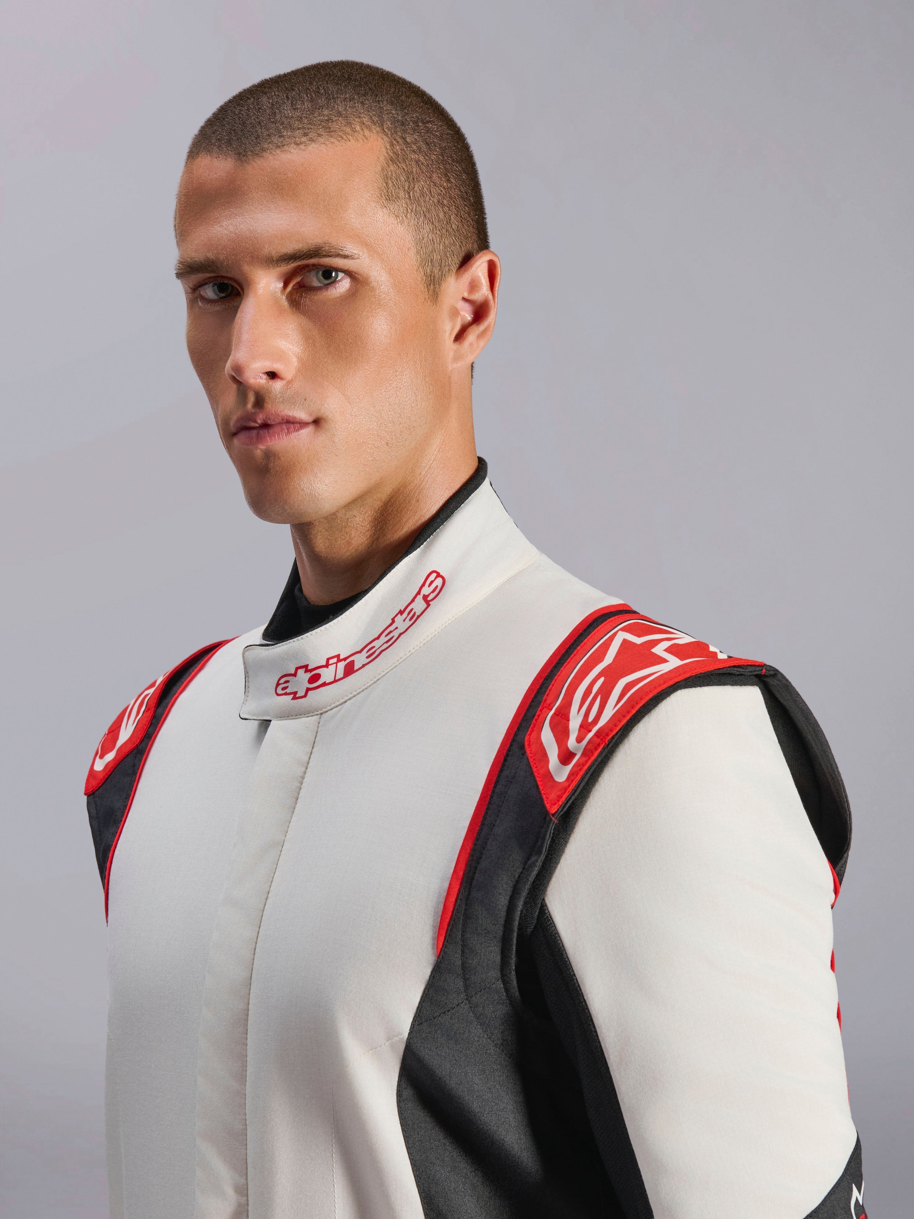 Kombinezon Alpinestars GP Tech V4, kombinezon samochodowy, biało-czarno-czerwony, prezentowany przez modela, czerwone panele na ramionach z logo Alpinestars, biały tors z czarnymi panelami bocznymi, kołnierz z logo, profesjonalny sprzęt do sportów motorowych