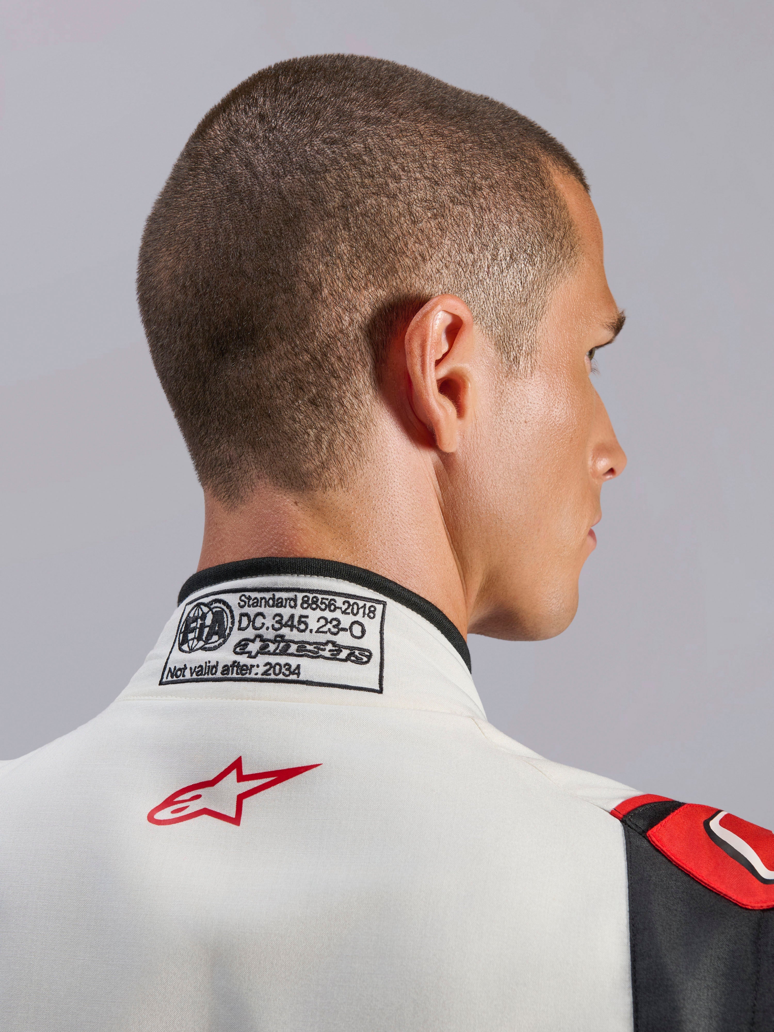 Kombinezon Alpinestars GP Tech V4, biało-czarno-czerwony samochodowy kombinezon wyścigowy, zbliżenie na tylną część kołnierza z etykietą certyfikacji FIA 8856-2018 i czerwonym logo gwiazdy, profesjonalny sprzęt ochronny do sportów motorowych
