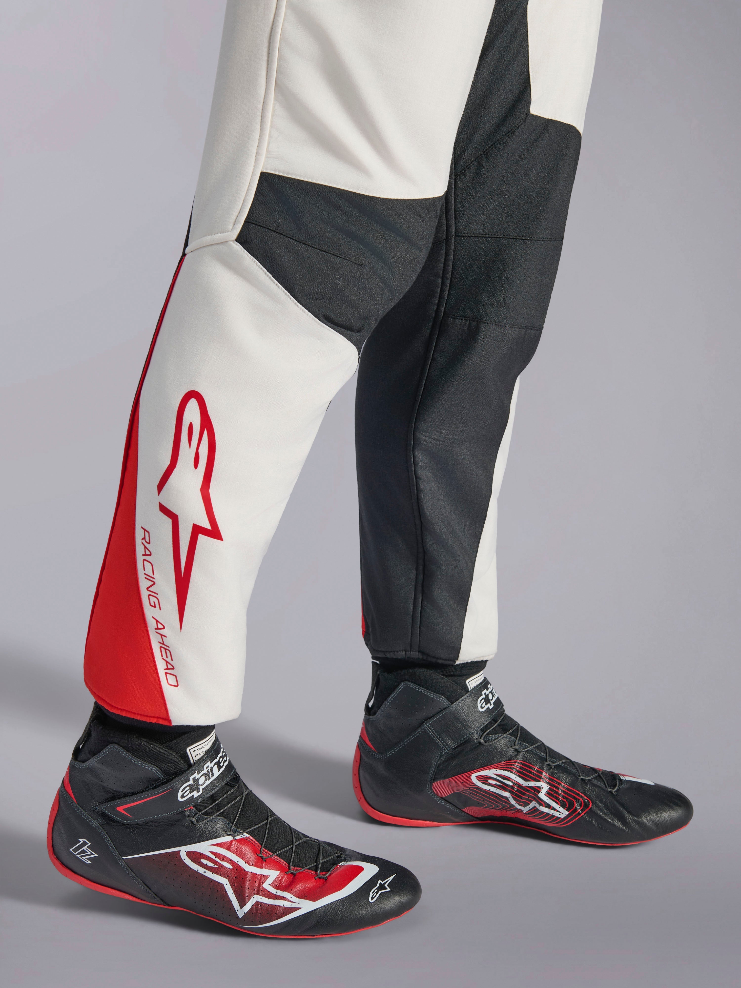 Kombinezon Alpinestars GP Tech V4, kombinezon samochodowy, biało-czarno-czerwony, widok na dolną część nogawek z białymi i czarnymi panelami, czerwonymi akcentami i napisem „RACING AHEAD”, noszony z czarnymi butami wyścigowymi, profesjonalna odzież do sportów motorowych