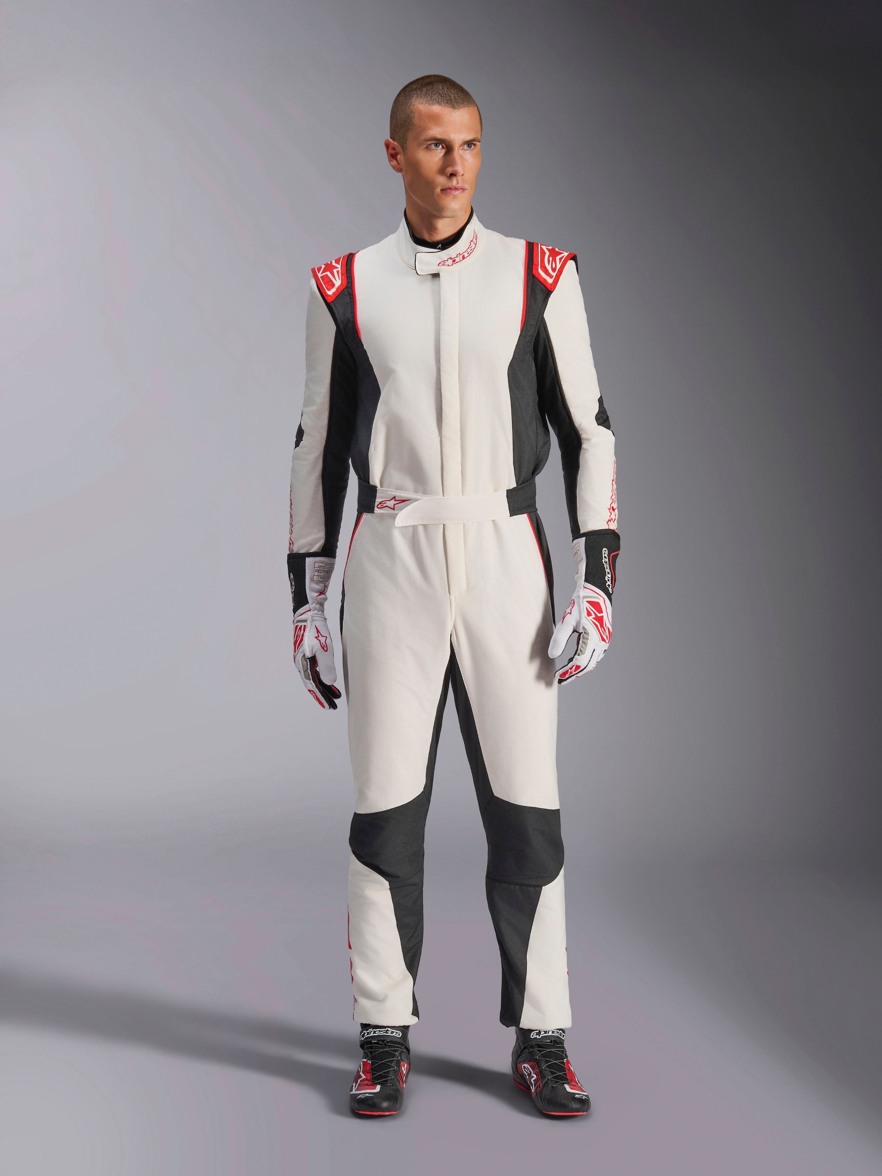 Alpinestars GP Tech V4 Suit, biało-czarno-czerwony jednoczęściowy kombinezon samochodowy, prezentowany przez modela z dopasowanymi rękawicami i butami wyścigowymi, z czerwonymi logo na ramionach i czarnymi panelami bocznymi, profesjonalny sprzęt do sportów motorowych przeznaczony na tor.