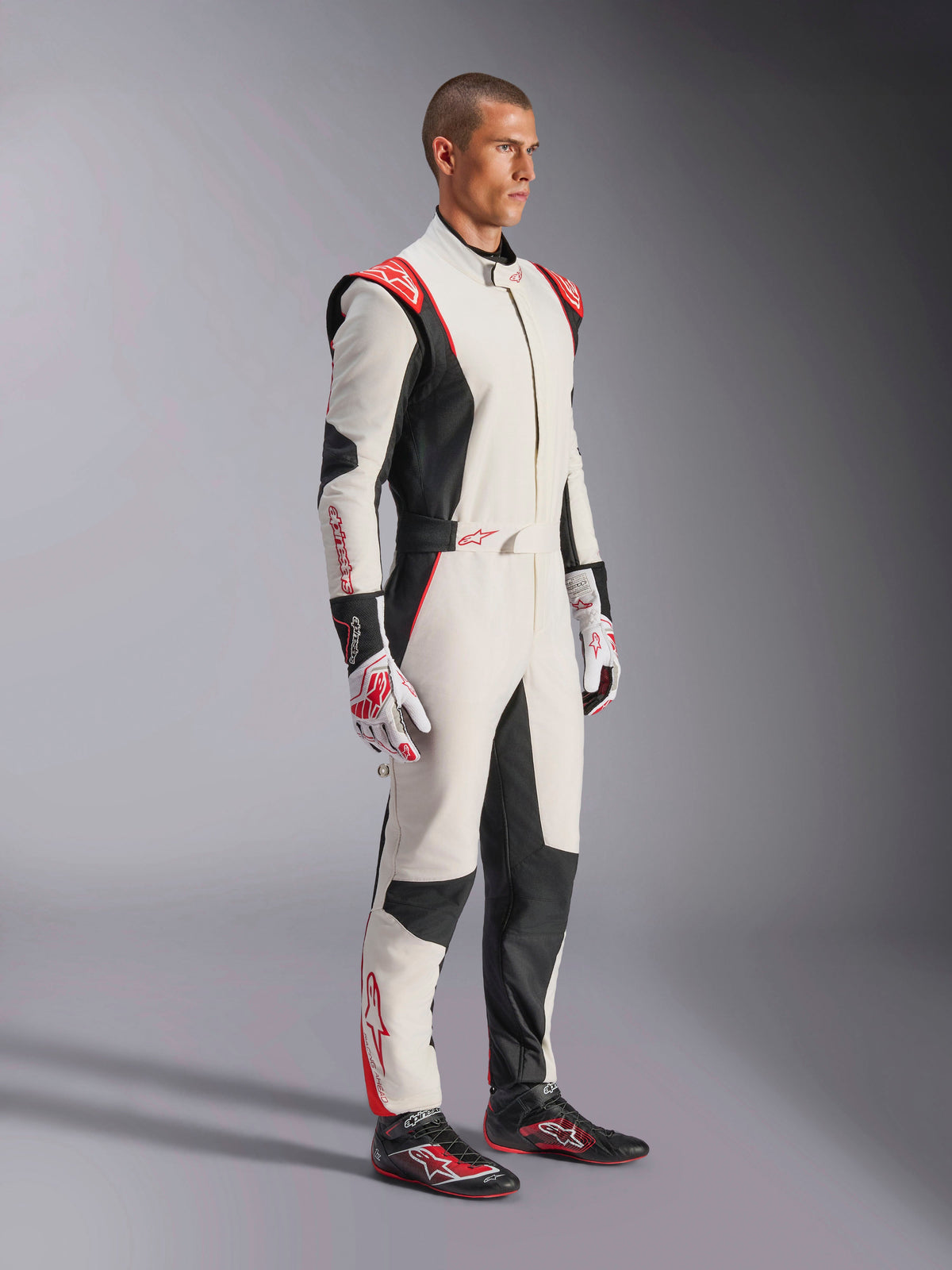 Kierowca wyścigowy w kombinezonie Alpinestars GP Tech V4, biało-czarno-czerwony, jednoczęściowy samochodowy kombinezon wyścigowy, pasujące rękawice i buty wyścigowe, profesjonalny sprzęt motorsportowy z logo Alpinestars i ergonomicznym designem
