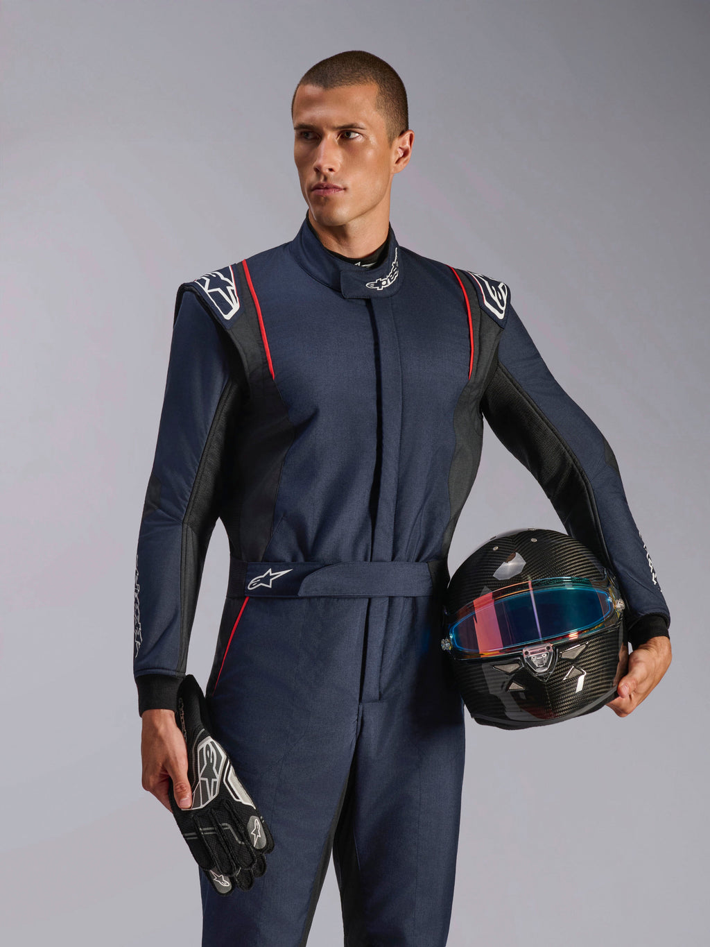 Model w kombinezonie Alpinestars GP Tech V4, granatowy kombinezon wyścigowy z czarnymi i czerwonymi akcentami, trzymający kask z włókna węglowego i rękawice wyścigowe, profesjonalna odzież ochronna do sportów motorowych