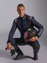 Kierowca wyścigowy w kombinezonie Alpinestars GP Tech V4, granatowy z czarnymi i czerwonymi akcentami, trzymający kask z włókna węglowego z iryzującym wizjerem, czarne rękawice i buty wyścigowe, profesjonalny samochodowy sprzęt wyścigowy