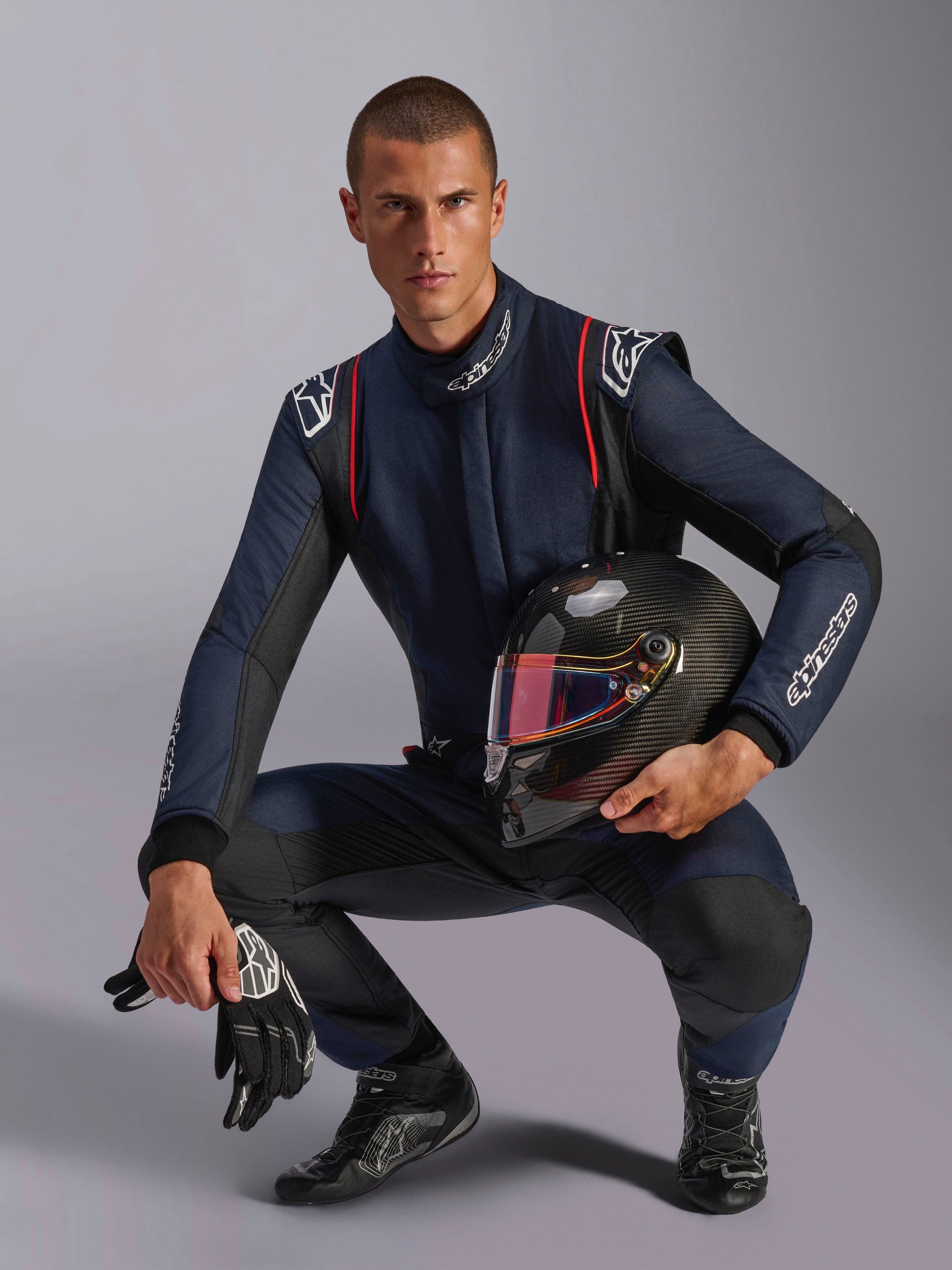 Kierowca wyścigowy w kombinezonie Alpinestars GP Tech V4, granatowy z czarnymi i czerwonymi akcentami, trzymający kask z włókna węglowego z iryzującym wizjerem, czarne rękawice i buty wyścigowe, profesjonalny samochodowy sprzęt wyścigowy