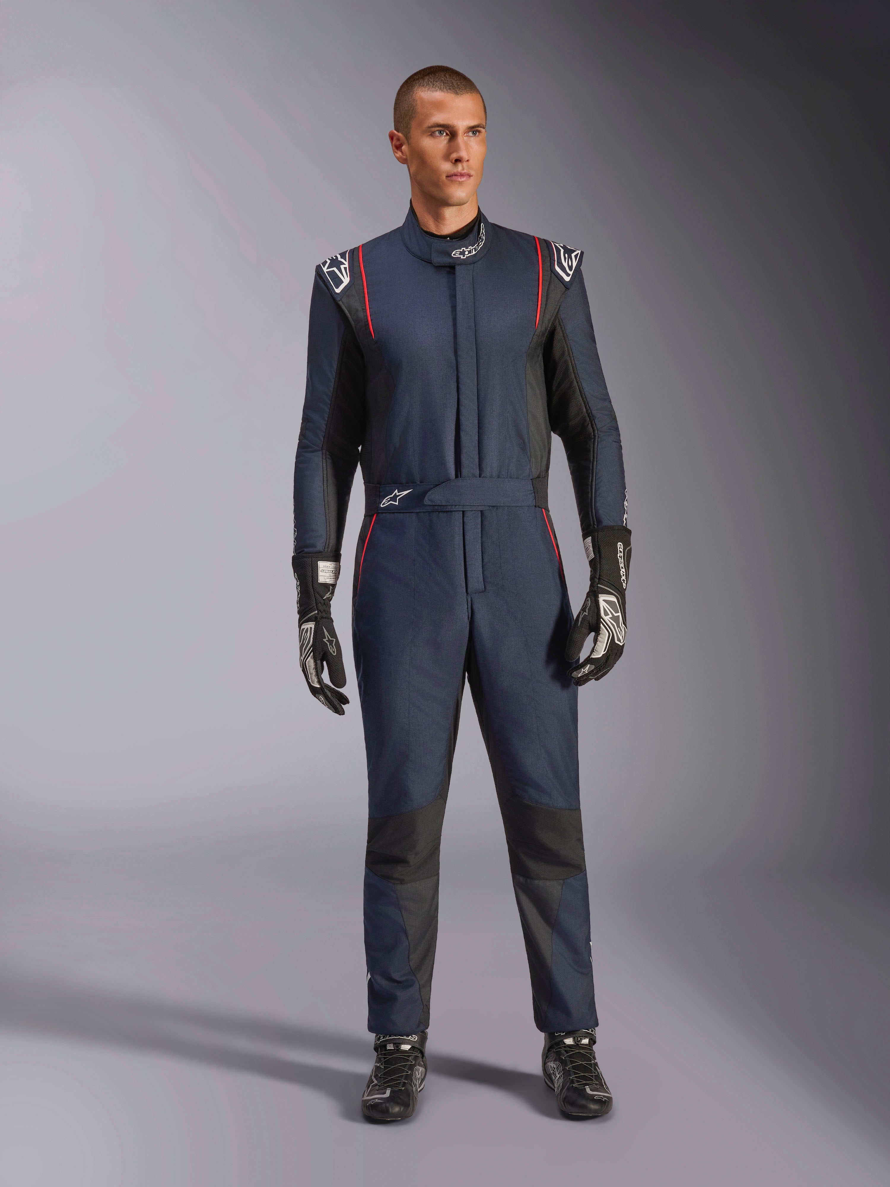 Kombinezon Alpinestars GP Tech V4, profesjonalny kombinezon wyścigowy w kolorze granatowym i czarnym z czerwonymi lamówkami, jednoczęściowy strój z certyfikatem FIA, noszony z czarnymi rękawicami i obuwiem wyścigowym, odzież dla kierowców sportów motorowych