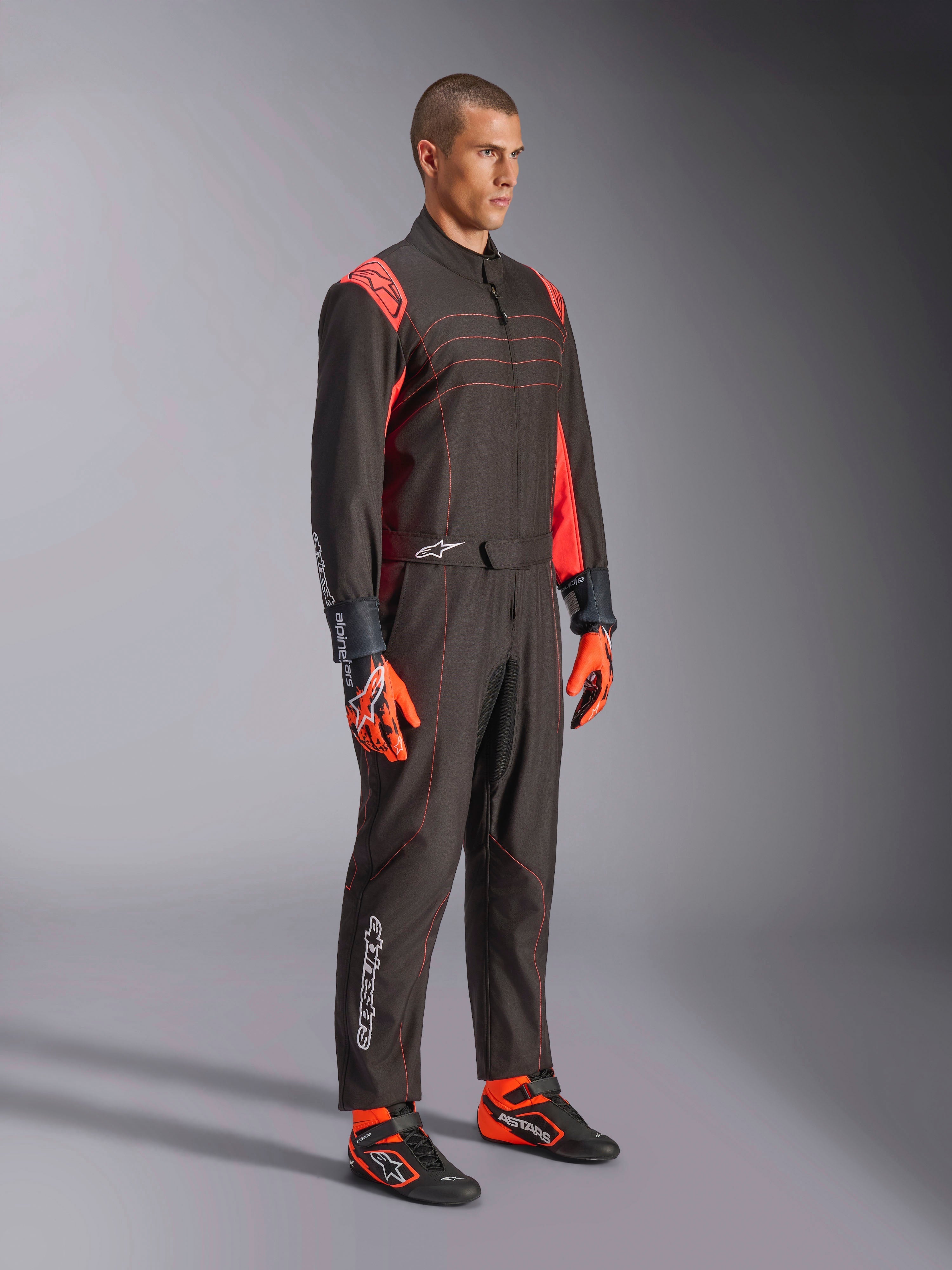 Alpinestars KMX-9 V3 Suit, kombinezon kartingowy, czarno-czerwony fluorescencyjny, prezentowany przez modela z pomarańczowymi rękawicami i czarnymi butami wyścigowymi, czerwone przeszycia, akcenty na ramionach i białe logo, sportowy design do profesjonalnych wyścigów