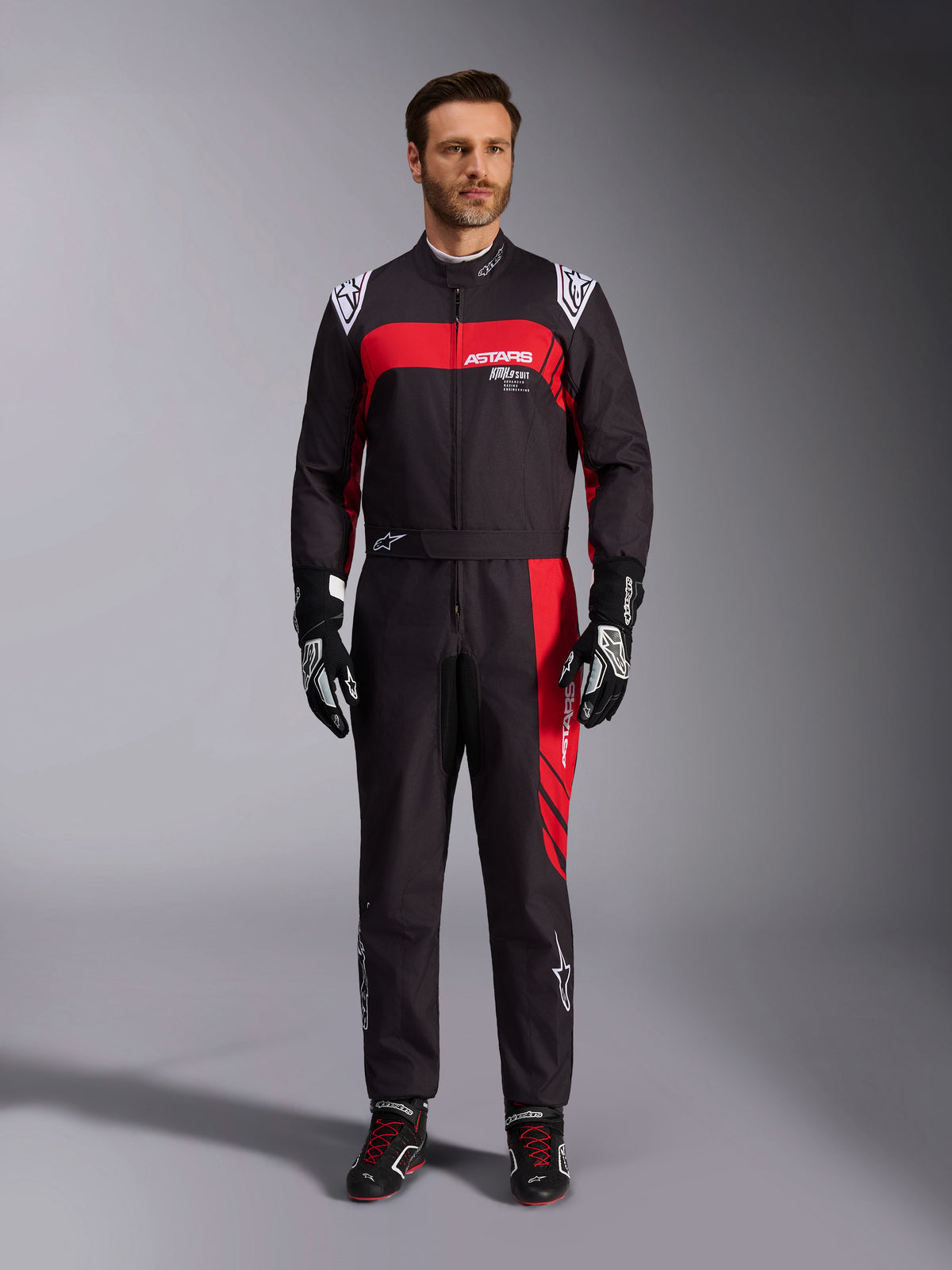 Alpinestars KMX-9 V3 Graphic 3 Suit, kombinezon kartingowy, czarno-czerwony, widok od przodu na modelu, wyraziste czerwone panele graficzne i białe logo, czarne rękawice i buty wyścigowe, profesjonalna odzież do sportów motorowych